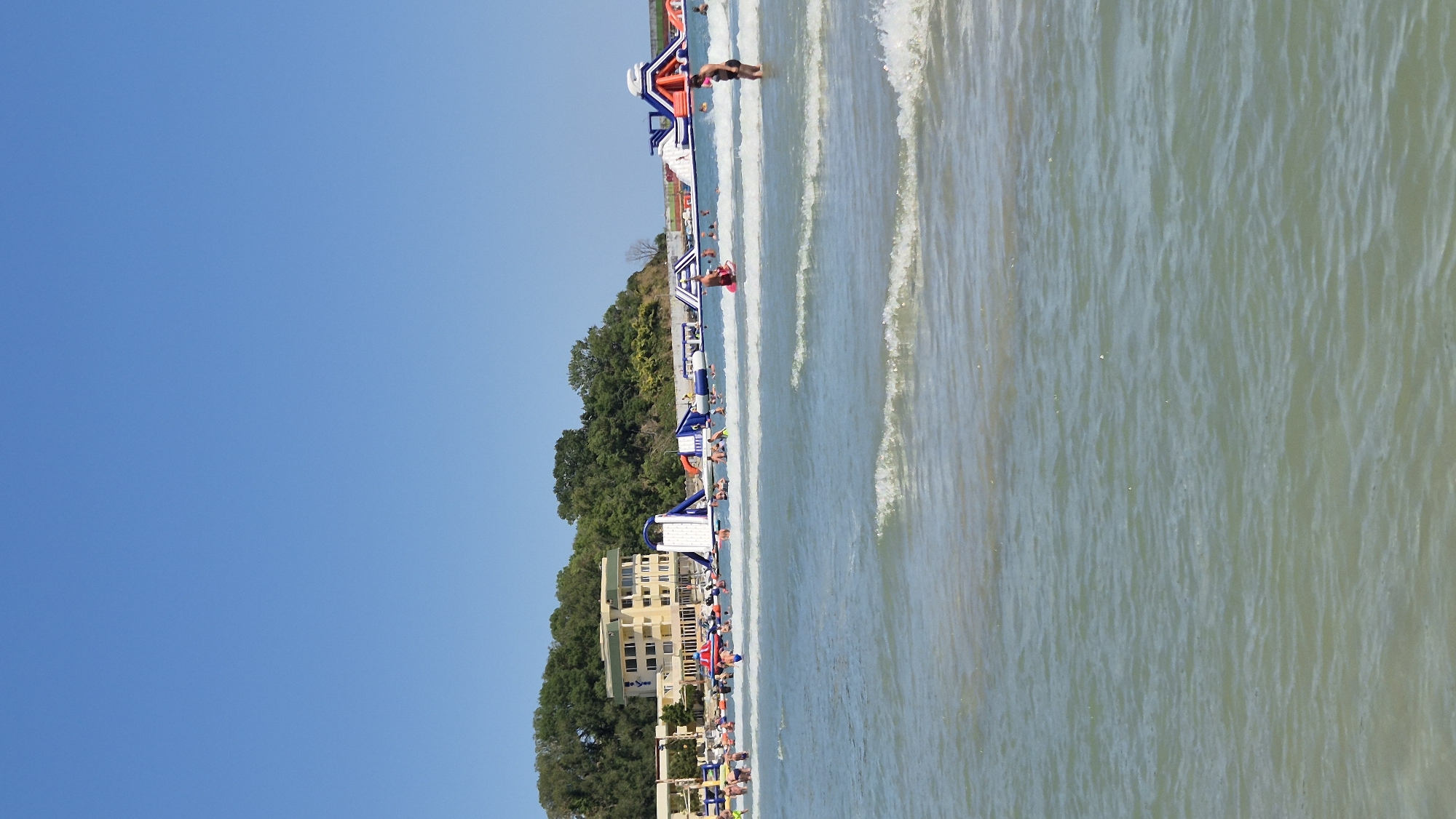 The image shows a sunny day at a beach in Kiten, Bulgaria, with the Black Sea in the foreground. The water is calm with gentle waves, and several people are swimming and enjoying the water. In the background, there are large inflatable water slides and obstacle courses set up in the sea, with more people interacting with them. A multi-story building, possibly a hotel, is visible on the shore, surrounded by lush green trees. The sky is clear and blue.