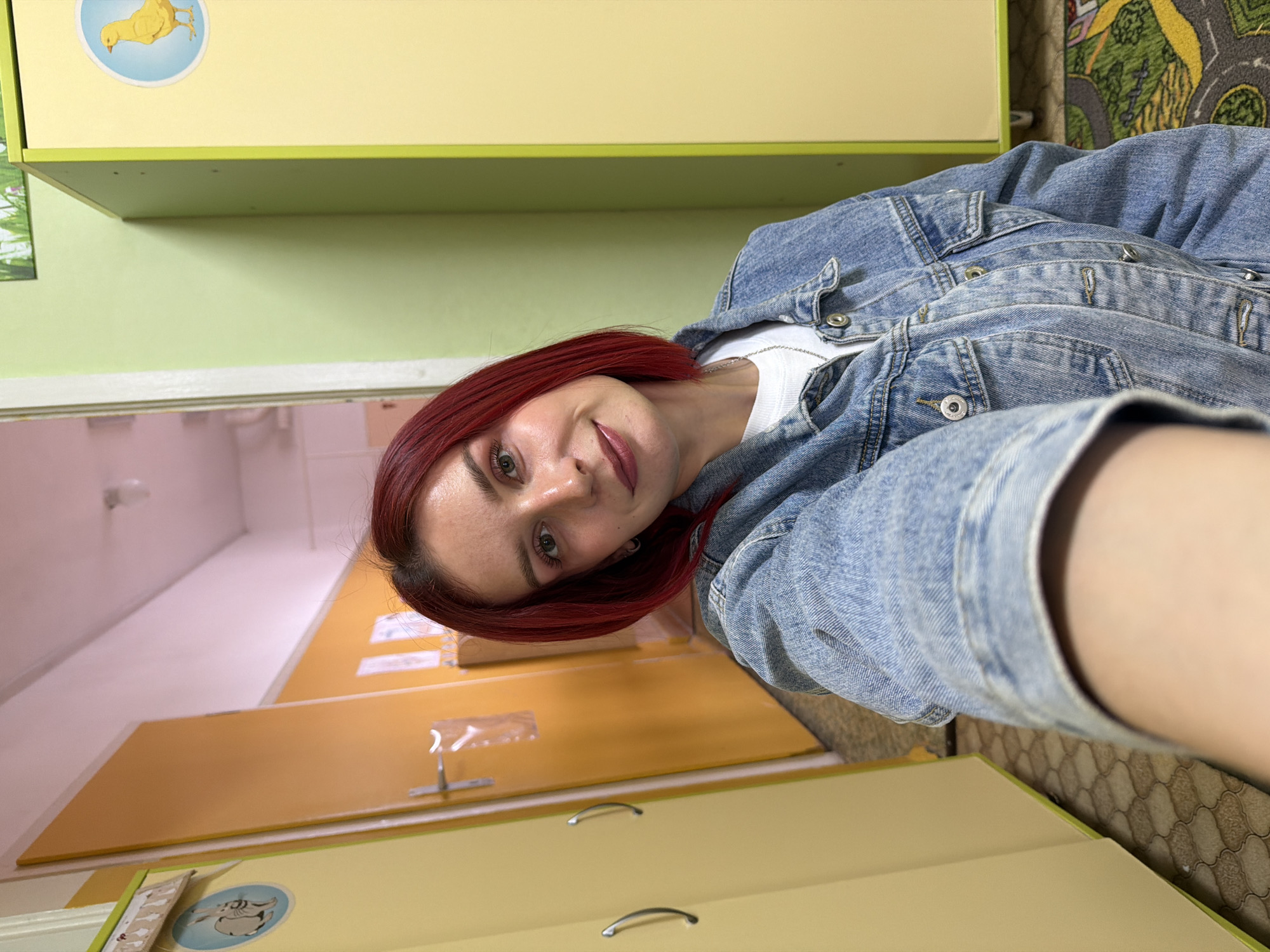 A woman with red hair and a denim jacket takes a selfie in what appears to be a hallway. There are light-yellow cabinets and beige doors. The location is in the city of Chaikovsky, Russia.