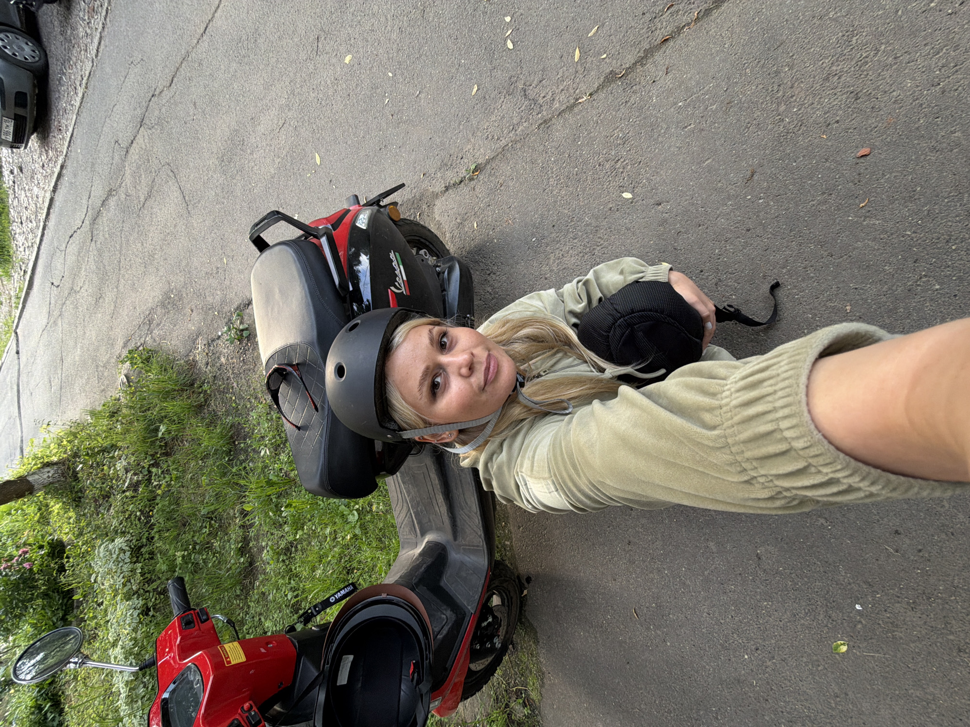 A young woman with blonde hair, wearing a light green hooded sweatshirt and black knee pads, is crouching on an asphalt road next to a red and black scooter. She is looking at the camera and holding a black helmet. The scooter has a black seat and a "Vespa" logo on the rear panel. The scene appears to be in an urban environment, with a sidewalk and grass area to the left of the road. A grey car is partially visible in the background, with a license plate that reads "Y986TM 18". The environment suggests it is daytime. Based on the license plate "Y986TM 18", this scene is likely located in the city of Sarapul, Russia, as the "18" region code corresponds to the Udmurt Republic, where Sarapul is located.