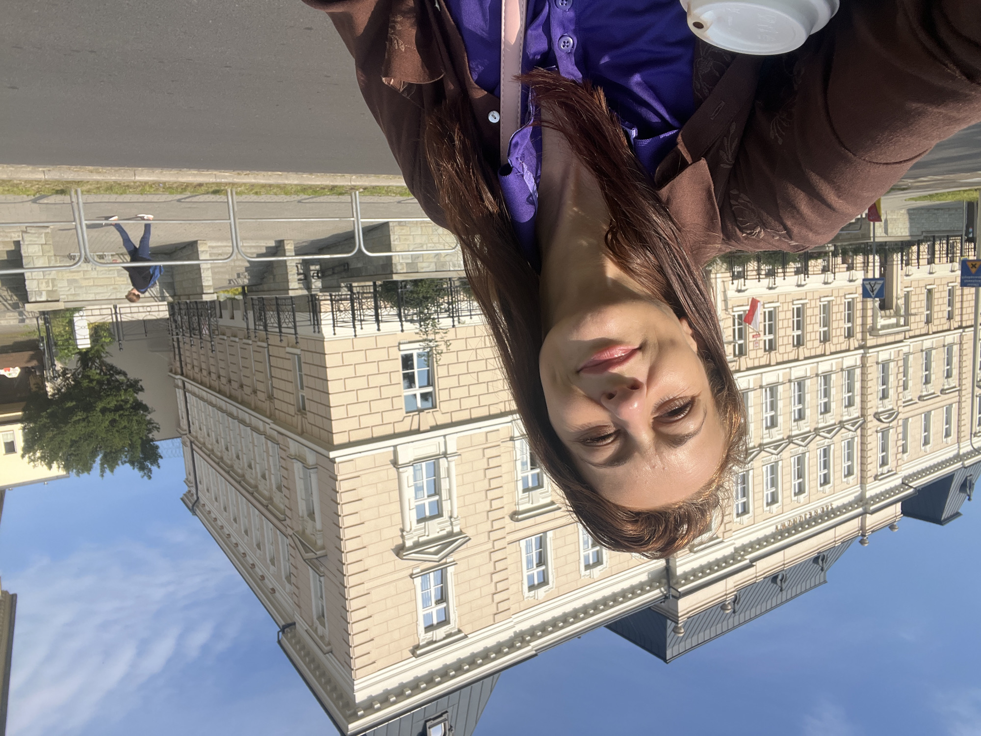 A medium shot captures a woman in the foreground, holding a white paper cup in her left hand. She is looking towards the camera with a slight smile, her face illuminated by sunlight. She has long, brown hair and is wearing a brown jacket over a purple collared shirt. The background features a large, ornate building with a light-colored facade, multiple windows, and a dark-tiled roof. The building appears to be in a city, with a street and sidewalk visible in front of it. A Polish flag is displayed on the building. Further back, a person with a blue backpack is walking away from the camera on the sidewalk. The sky is clear and blue with some scattered clouds.