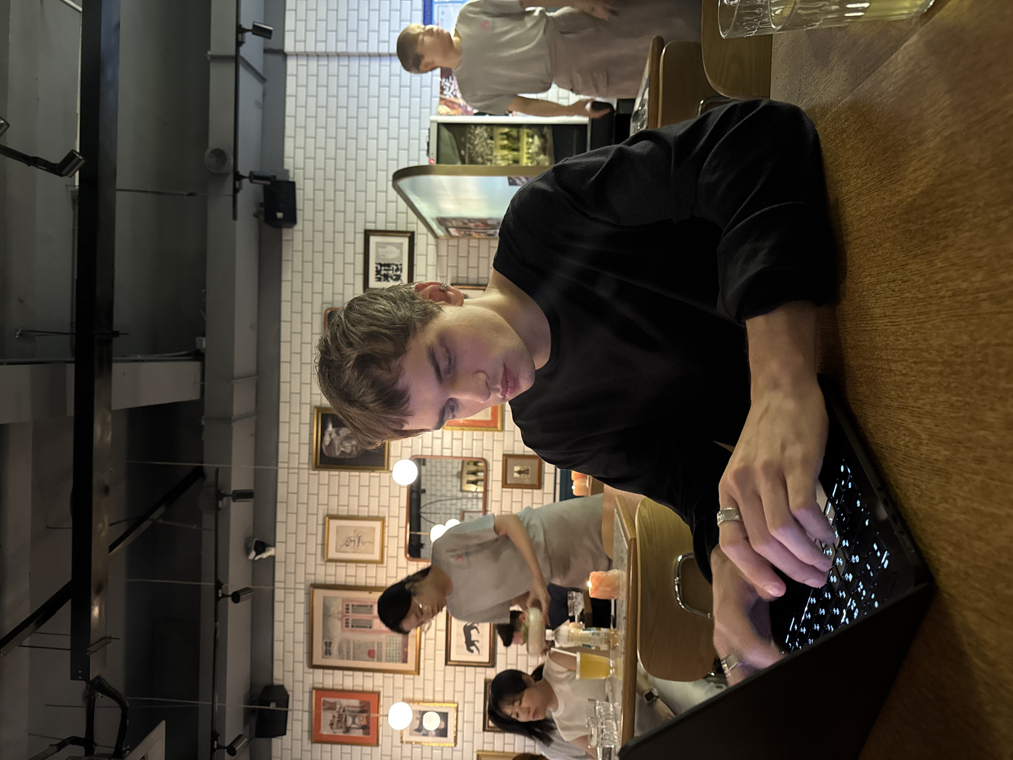 The image depicts a young man with short, light brown hair, wearing a black long-sleeved shirt and several rings on his fingers, seated at a wooden table in what appears to be a restaurant in Xuhui District, China. He is actively typing on a black laptop, which is positioned on the table in front of him. The background reveals a dimly lit restaurant interior with exposed ceiling elements and industrial lighting fixtures. White tiled walls are adorned with framed pictures and artwork. Several other individuals are present in the background: a woman in a light gray uniform is carrying a bowl, suggesting she might be a server; another woman with dark hair is seated at a table to the left, looking down; and a man in a gray shirt and apron is standing further in the background near what looks like a bar area. Glasses and a lit candle are visible on the table in the foreground. The overall atmosphere suggests a casual dining setting with patrons engaged in various activities.