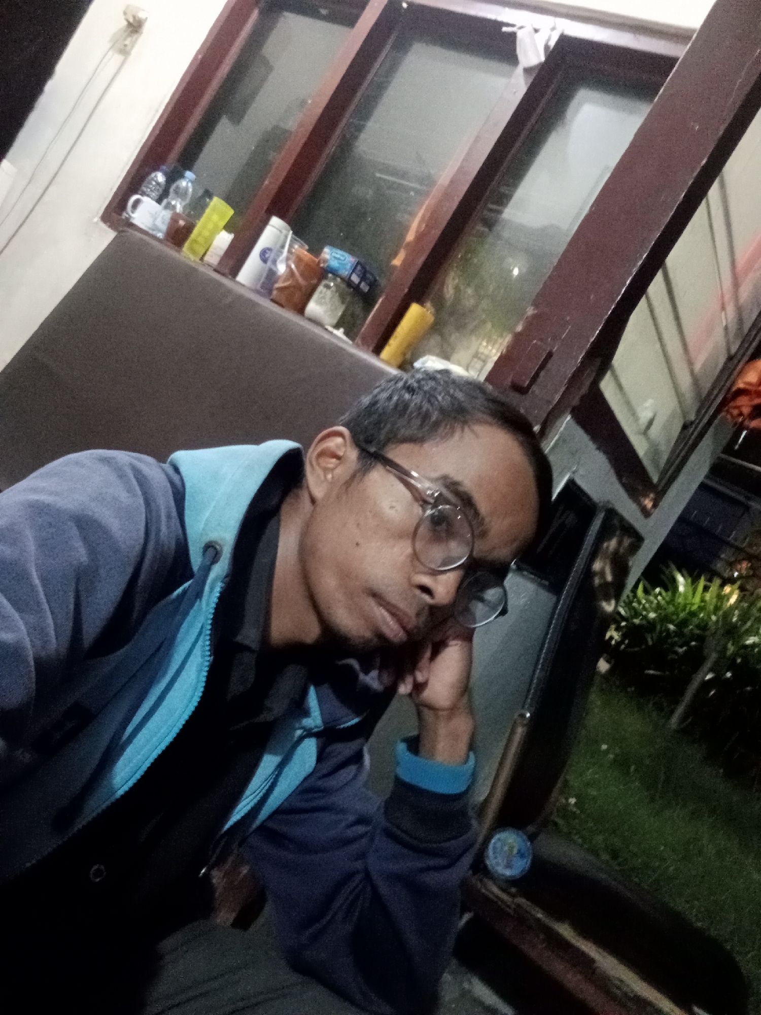 The image captures a selfie taken by a man with glasses, wearing a blue hooded jacket over a black shirt. He appears to be seated indoors, with a dark brown couch or chair in the foreground. Behind him, a window with wooden frames is visible, and on the windowsill, several items including bottles, a mug, and containers are arranged. The background outside the window shows greenery, suggesting an outdoor area with plants. The lighting in the image is dim, indicating it was taken at night or in low-light conditions. The man's facial expression is thoughtful, with his head resting on his hand. The scene appears to be in a residential or a casual setting within the city of Surabaya, Indonesia, judging by the common visual cues of Indonesian architecture and surroundings.