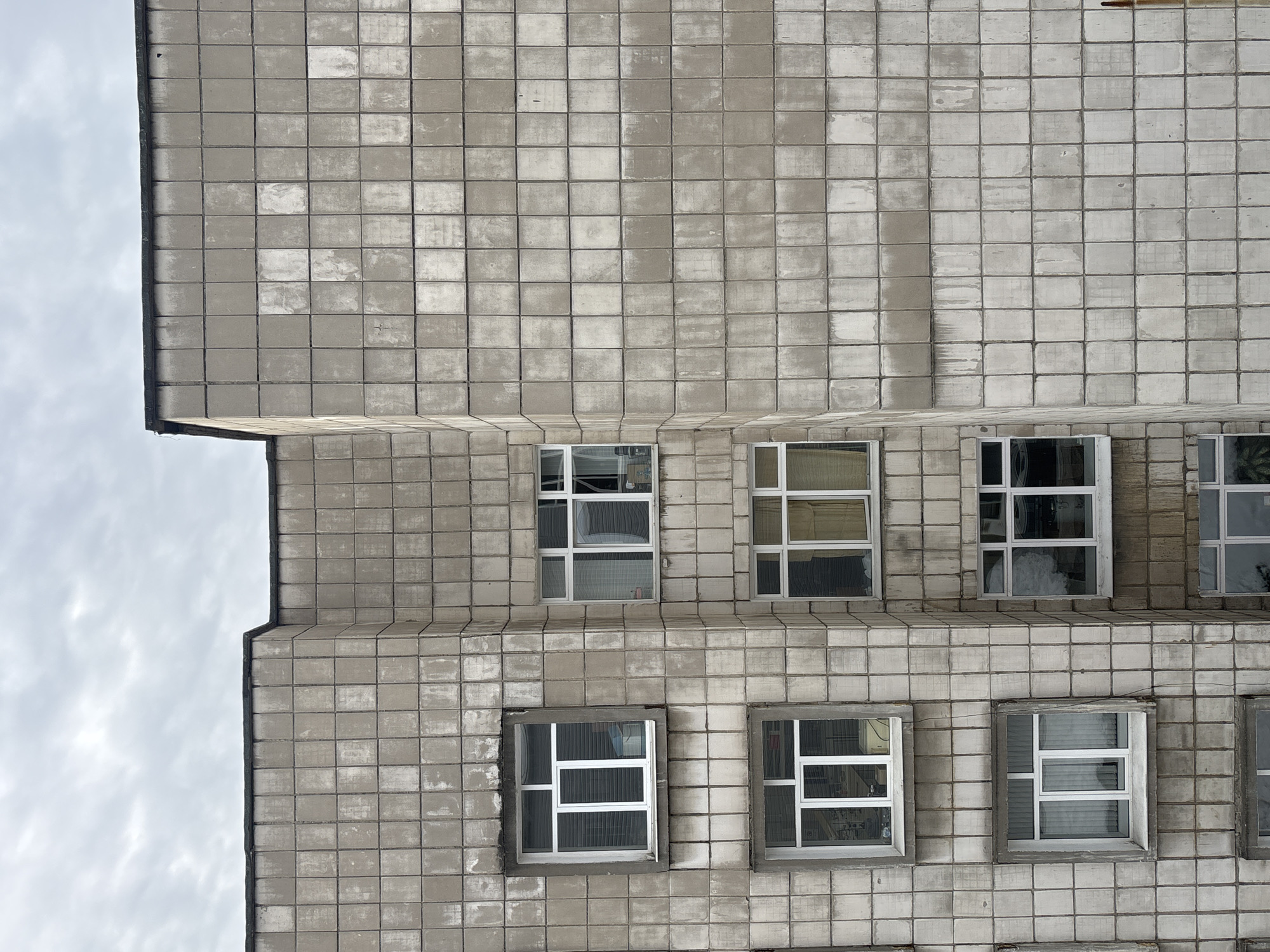 A low-angle, medium shot depicts the exterior wall of a building with a repetitive square tile facade, likely located in Perm, Russia. The building features several rectangular windows with white frames and multiple panes. The windows reflect the sky and interior elements. The tilework shows signs of weathering. The upper left corner of the frame reveals a glimpse of overcast sky.