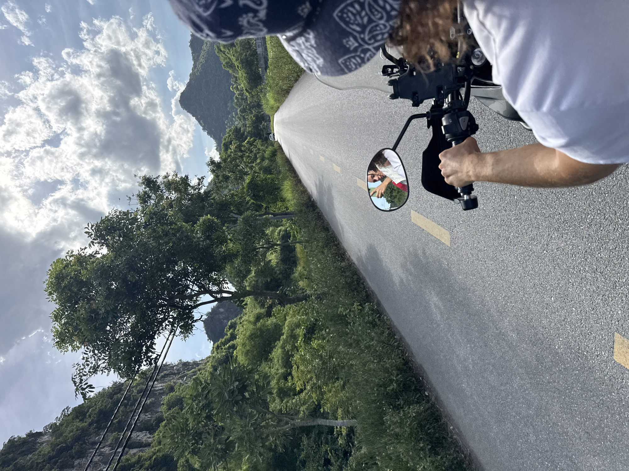 The image captures a perspective from someone riding a motorcycle on a paved road in Nam Hoa Lư Ward, Vietnam. The road stretches forward, flanked by lush green vegetation and imposing karst mountains. In the foreground, the rider's arm and shoulder are visible, along with the handlebars of the motorcycle. A rearview mirror reflects two individuals, one holding a smartphone. The sky above is partly cloudy, with sunlight breaking through. The overall scene depicts a scenic journey through a rural Vietnamese landscape.
