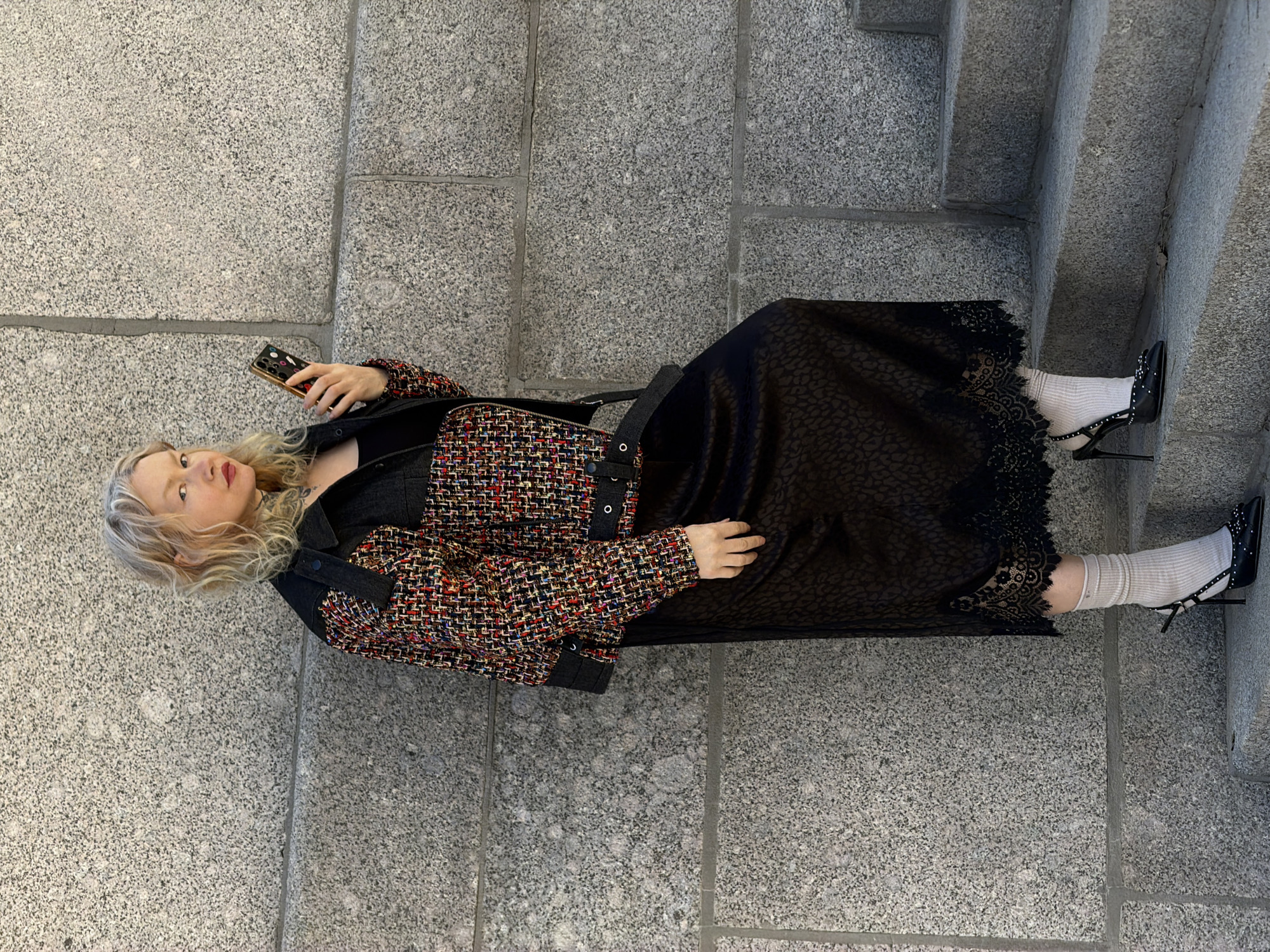 A woman with blonde hair stands on a set of stone steps adjacent to a granite wall in Saint Petersburg, Russia. She is holding a cell phone in her left hand and is dressed in a multi-colored tweed jacket over a black dress with lace trim, white socks, and black high-heeled shoes. She also has a black purse slung over her shoulder. The granite wall is made up of large rectangular blocks.