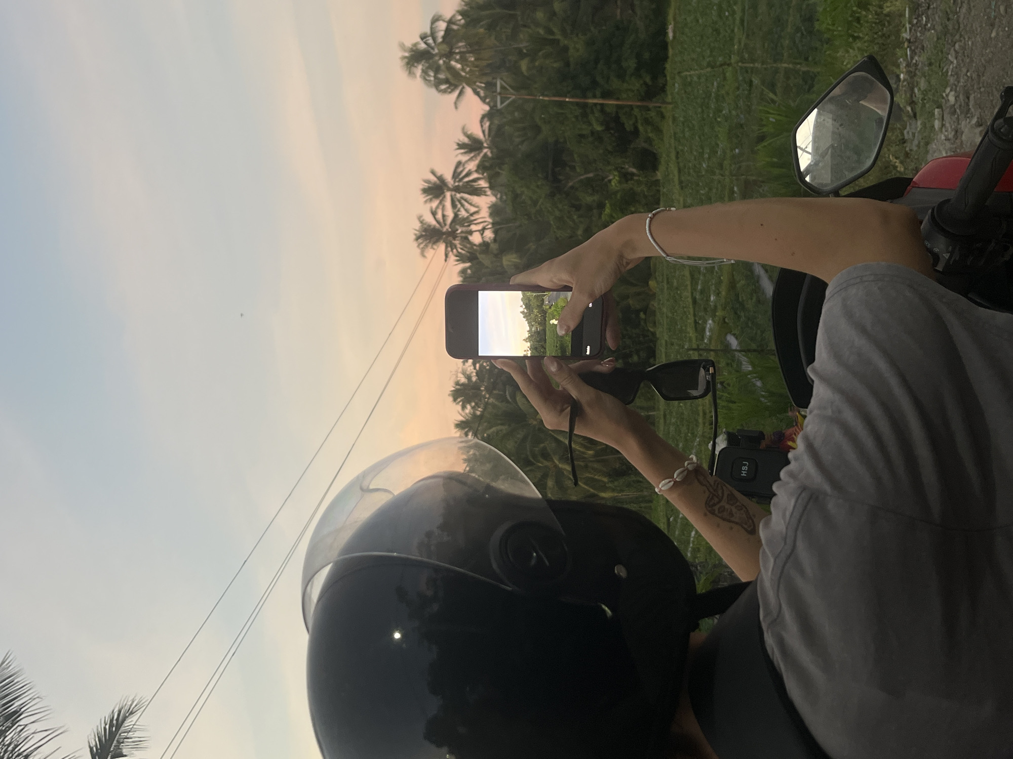 A person wearing a black helmet and a grey t-shirt is seated on a red scooter, holding a smartphone to capture a scenic view. The person's left arm is adorned with a bracelet of white shells and a tattoo of a mushroom on their forearm. The smartphone is displaying a landscape with a road and a field, framed by lush green trees and palm trees under a soft pink and blue sky, indicative of a sunset. The background features a rural landscape with agricultural fields and utility poles. This scene is set in Loloan, Indonesia.
