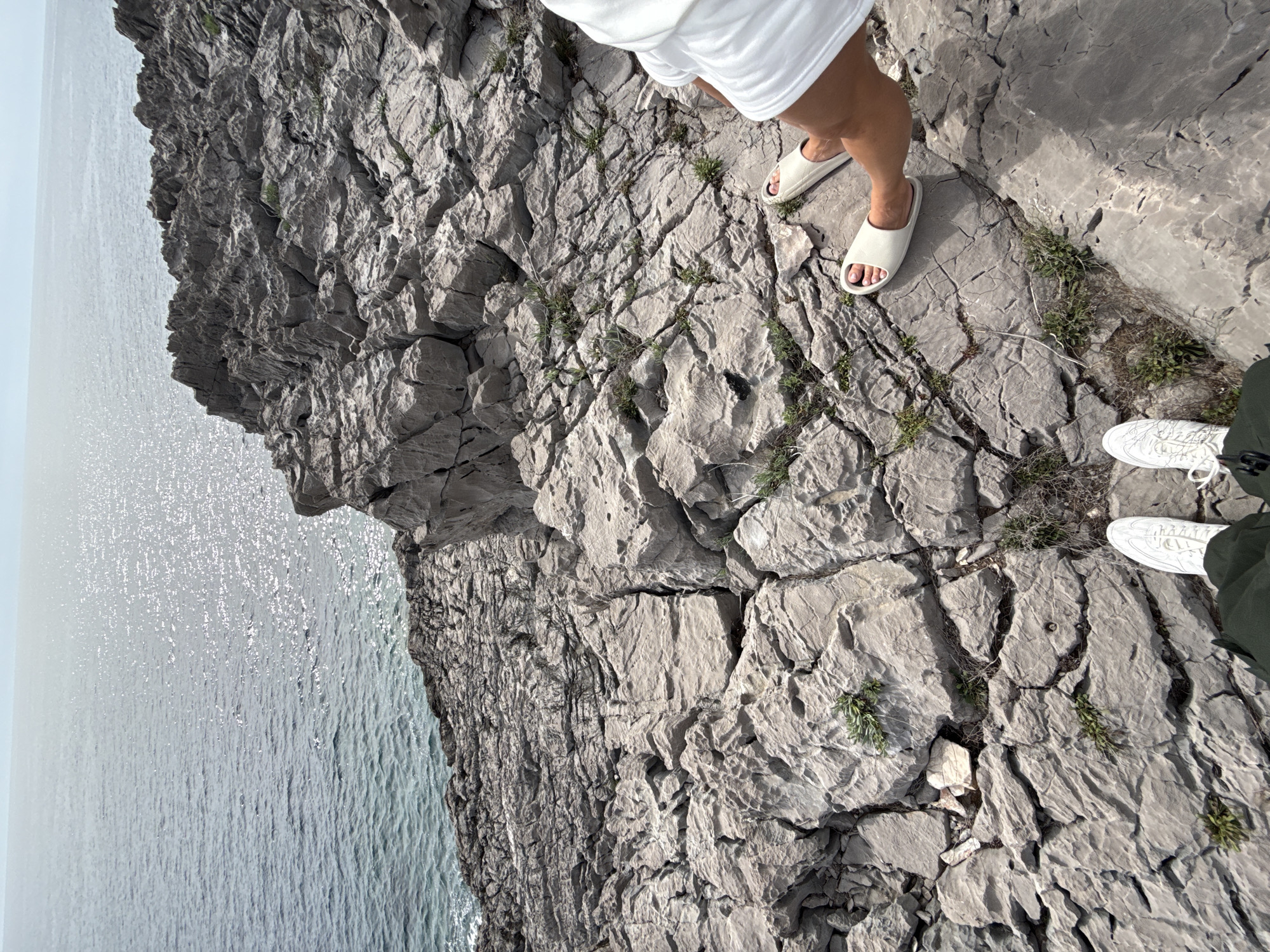 The image captures a view from a rocky cliff in Boreti, Montenegro. To the left, the Adriatic Sea stretches out, its surface reflecting sunlight. The cliff face is composed of gray rock formations, with sparse patches of vegetation growing in the cracks. Two individuals are visible: one is wearing white slides and a white outfit and the other wearing white sneakers.