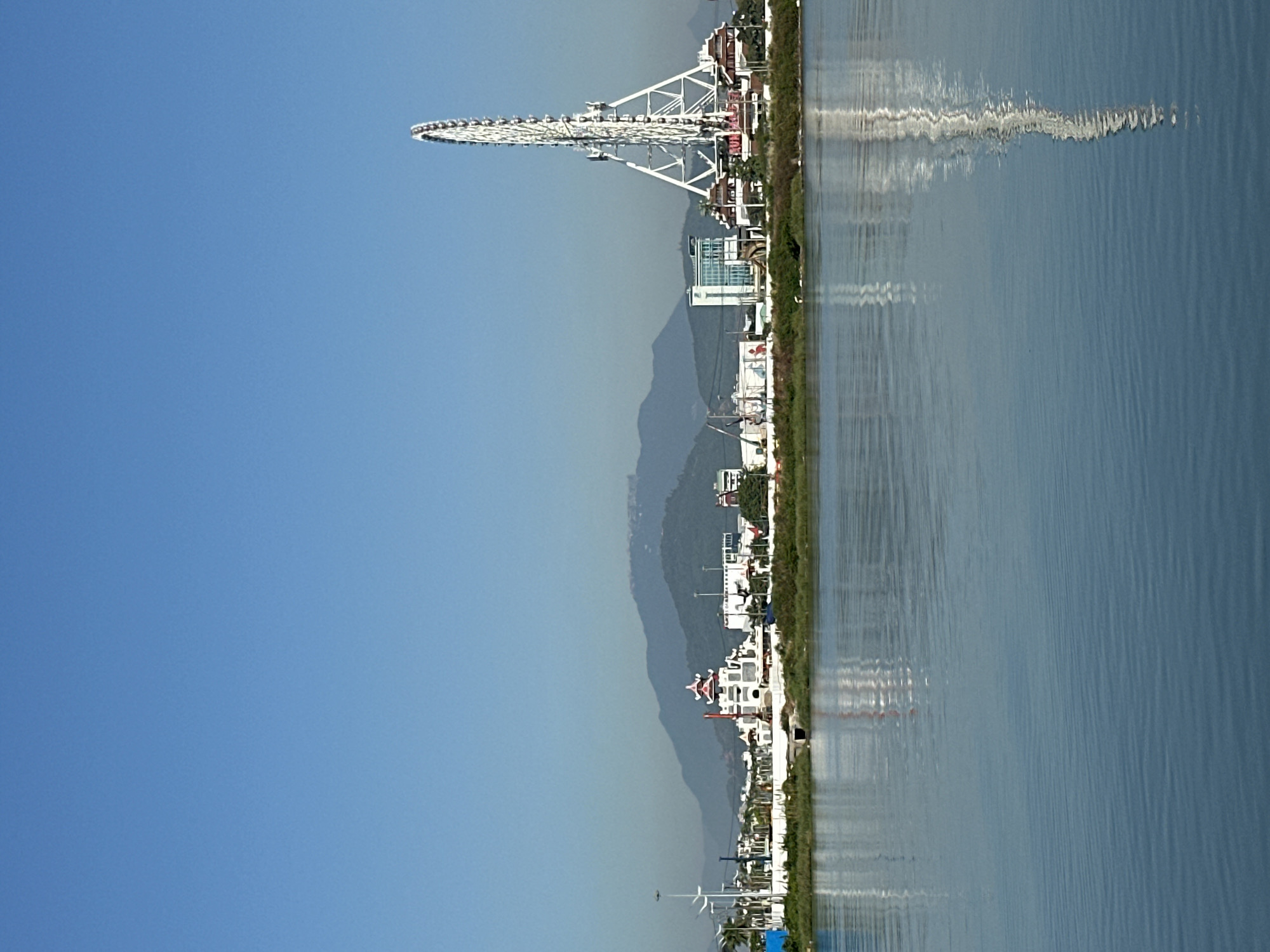 The image captures a waterfront scene in the city of Da Nang, Vietnam, dominated by a large white Ferris wheel on the right side of the frame. The Ferris wheel is tall and has a prominent triangular support structure. In the background, a range of lush green mountains forms a natural backdrop. A body of calm water occupies the lower half of the image, reflecting the sky and the structures along the shore. Along the shoreline, there are several buildings, including a white structure with a traditional East Asian architectural style, featuring a red-tiled roof. Other modern buildings and some smaller structures are also visible. The sky is clear and blue. There are no people or animals readily apparent in the image. The scene appears to be a public area, possibly a park or entertainment district.