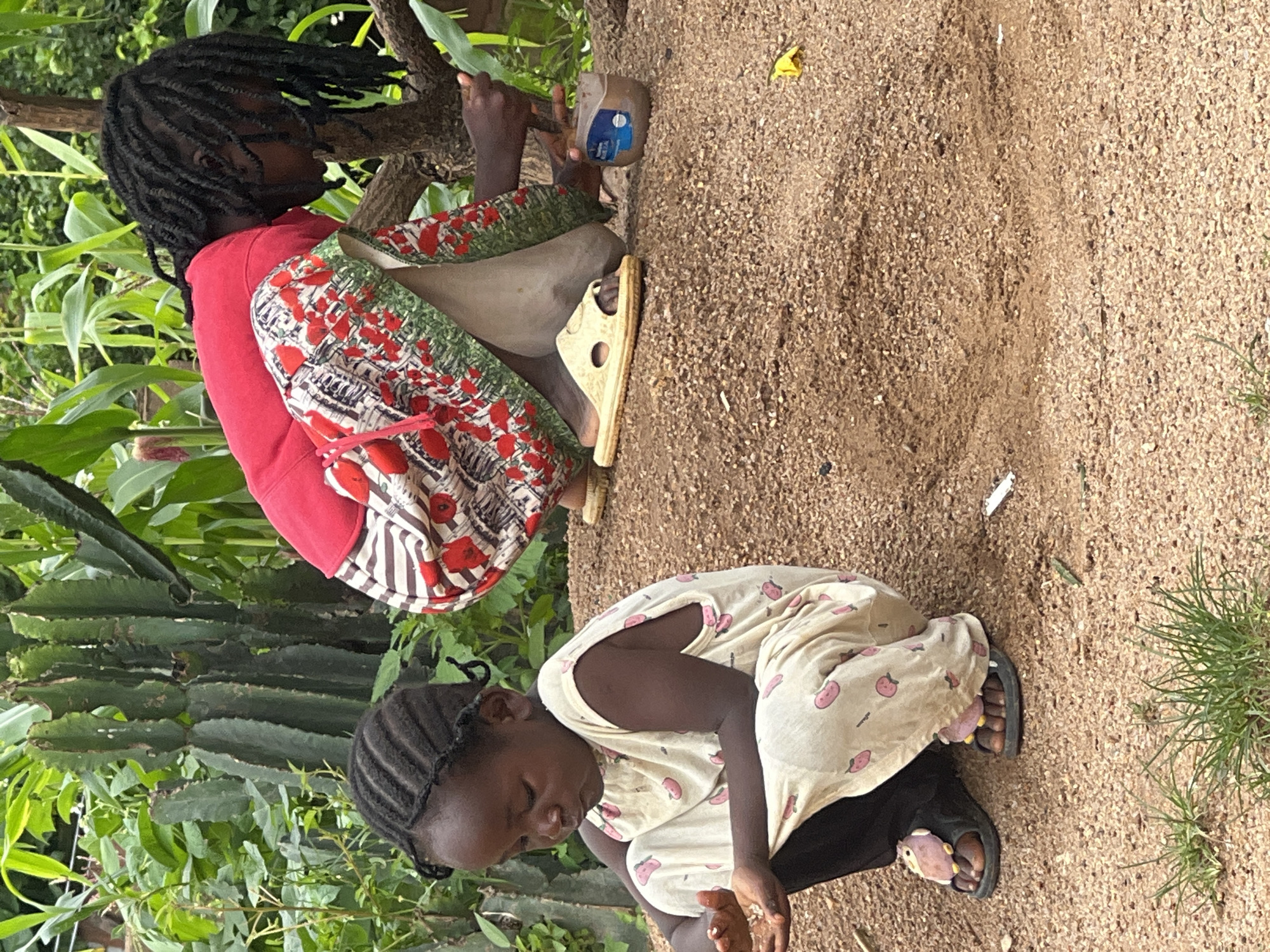 Two young children are in a dirt-covered outdoor area, possibly in a rural part of Jos, Nigeria, with lush green vegetation and corn stalks in the background. A younger child, dressed in a light-colored pajama-like outfit with pink fruit patterns, is crouched on the ground, looking down at their hands, which appear to be holding small white objects. This child is wearing black pants and sandals adorned with pink cartoon characters. An older child, wearing a red top and a patterned skirt with red floral designs, is also crouched nearby. They are reaching towards a small brown jar or container placed on the ground, holding a green leaf or twig in their right hand. The older child has dark, braided hair and is wearing light-colored slide sandals. The ground is covered in coarse sand and small pebbles.