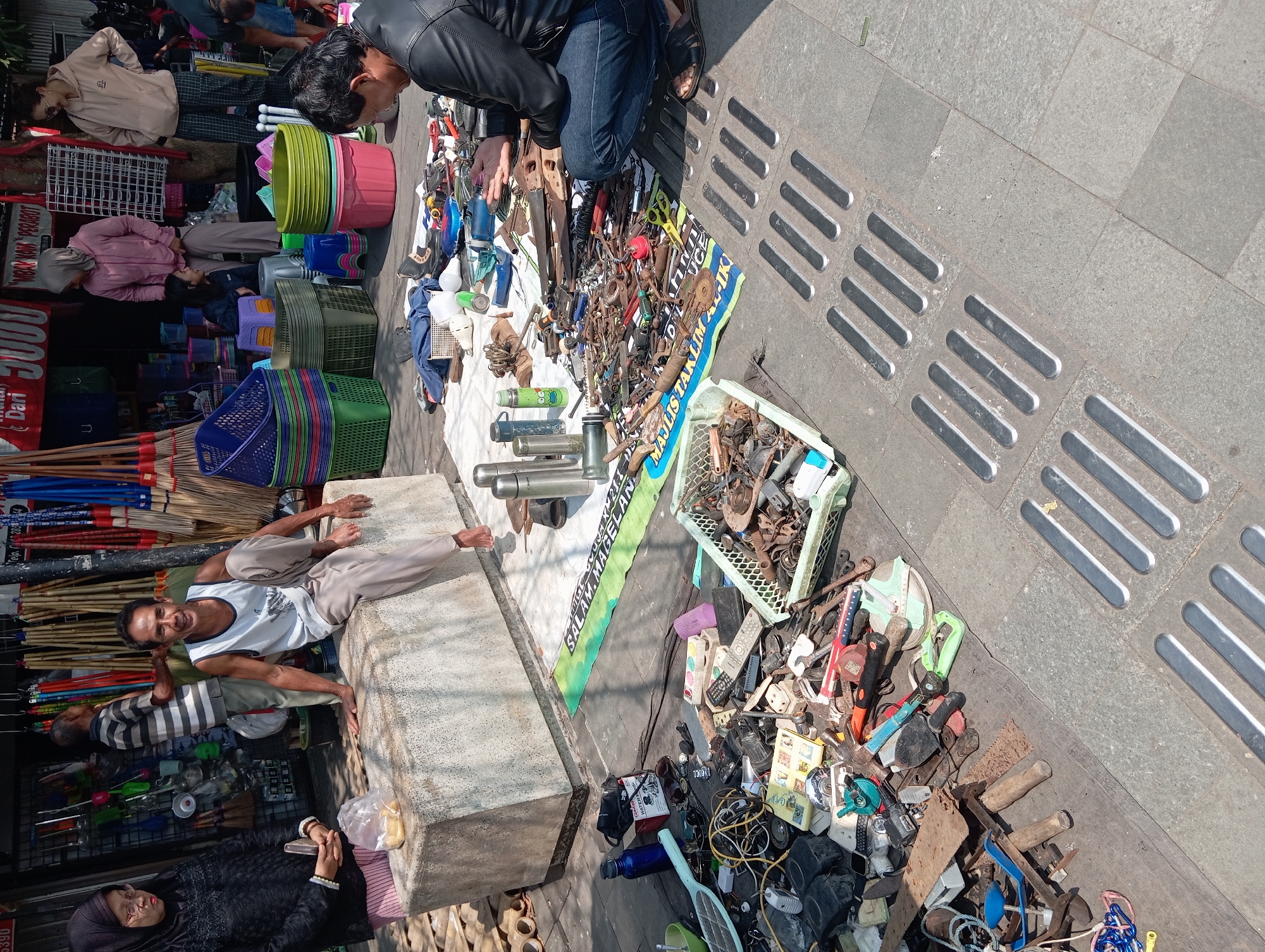 A street vendor is selling various items on the sidewalk in front of shops in Borobudur, Indonesia. The scene is bathed in daylight. A man in a white tank top and beige pants sits on a concrete block to the left, looking at the camera and smiling. To his right, a man wearing a black leather jacket and blue jeans crouches down, examining items spread on a blue and green mat. More items, including tools, electronics, and household goods, are scattered on the ground to the left and in baskets. Behind the vendor on the block, a woman in a black hijab and glasses stands holding a phone and a plastic bag of food. In the background, other shoppers and vendors are visible, as well as stacks of colorful plastic baskets and brooms in front of the shops. The ground is paved with stone, with tactile paving strips for the visually impaired.