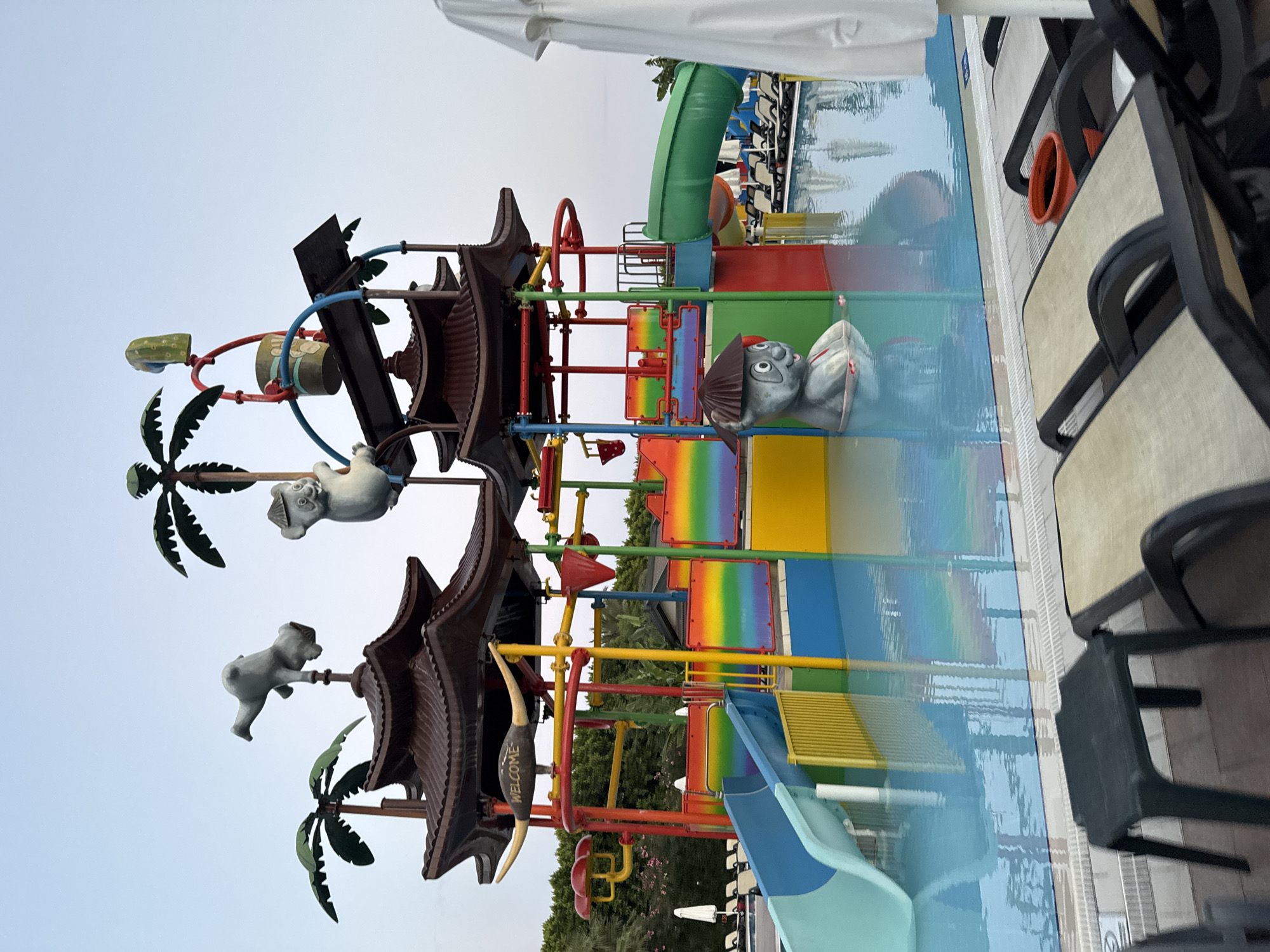 The image shows a water park in Aksu, Turkey. The water park features a multi-level structure with colorful slides, platforms, and decorative elements, including two gray cartoonish figures that appear to be animals. There are palm trees and lush green trees in the background, suggesting a warm climate. The water in the pool is clear blue. In the foreground, there are lounge chairs and a small plastic table by the poolside. The sky is overcast. The overall scene depicts a recreational area designed for water-based activities.