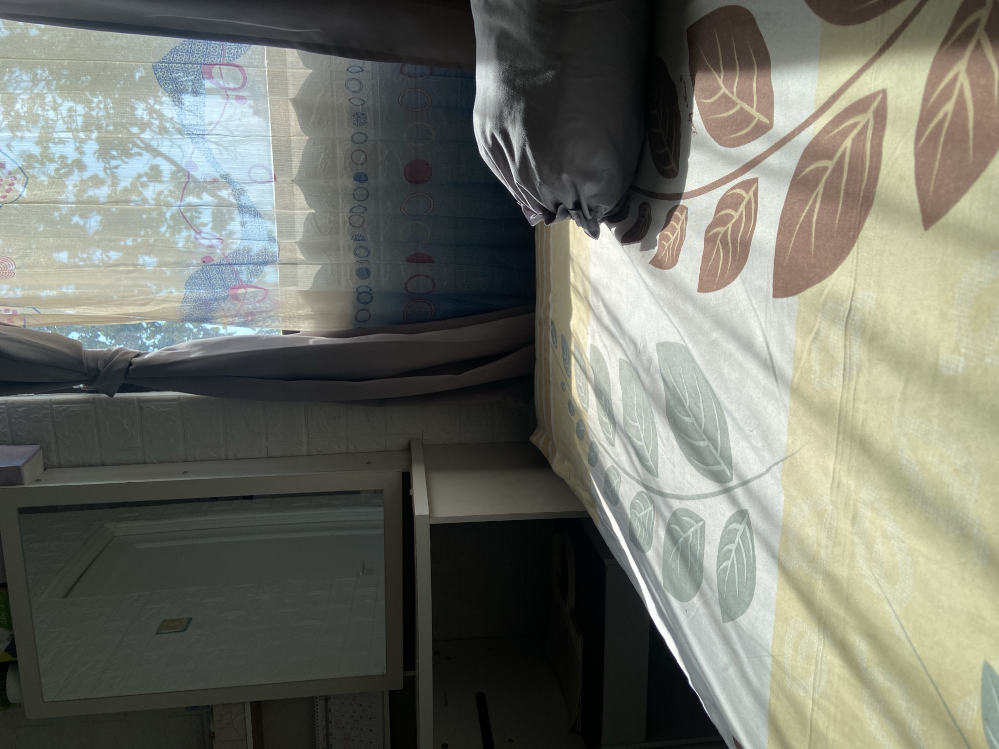 In Margorejo, Indonesia, a bedroom scene unfolds with natural light streaming through a window, casting shadows across a bed. The bed is covered with a comforter featuring a leaf pattern in shades of yellow, green, and brown. A gray bolster pillow rests on the bed, partially obscuring the comforter. To the left, a vanity with a mirror is visible, its surface reflecting the room. The wall behind the vanity and bed is textured with what appears to be a white brick pattern. The window is dressed with sheer, patterned curtains that diffuse the sunlight, revealing the silhouette of trees outside. The overall impression is one of a quiet, sunlit morning in a residential setting.