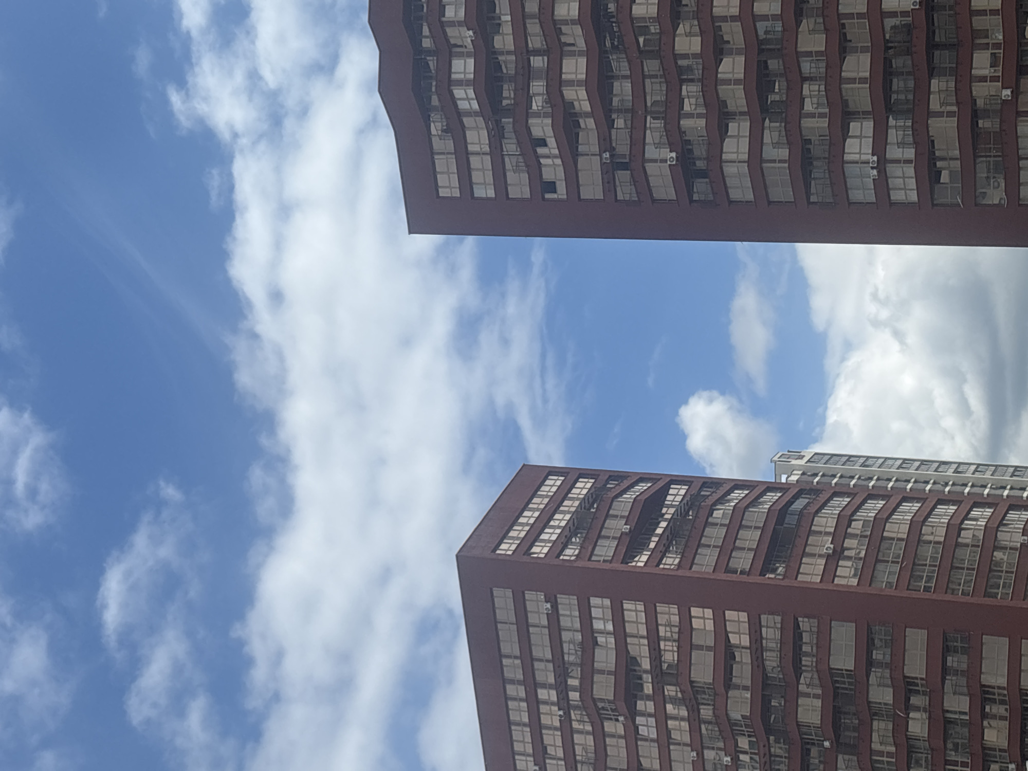 The image shows a view of two modern high-rise buildings against a blue sky with scattered white clouds. The building on the left is primarily visible, with its dark red facade featuring numerous rectangular windows arranged in a grid pattern. Balconies with curved railings are also visible, interspersed between some of the window levels. In the background, a portion of another, lighter-colored building can be seen to the right of the main red building. The building on the far right of the frame is also a high-rise with a dark red facade and a similar window and balcony design to the building on the left. The perspective is from below, looking upwards at the structures. This scene is located in the city of Minsk, Belarus, specifically in the area of the Minsk World complex.
