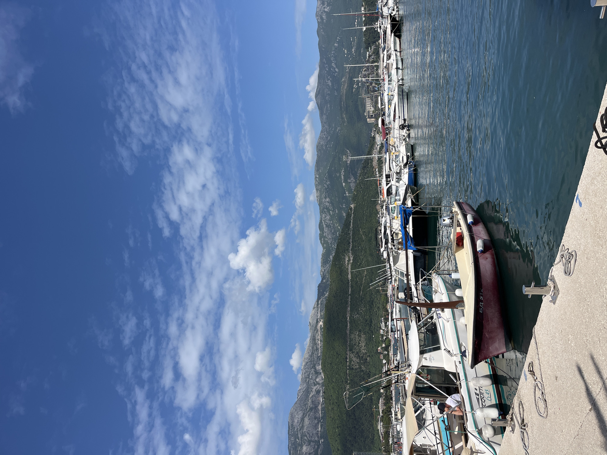 The image depicts a marina in Budva, Montenegro, featuring numerous yachts and boats docked along a concrete pier. The foreground shows several boats closely moored, with a mix of maroon, white, and blue colors. Clear calm water reflects the masts and superstructures of the vessels. The background showcases a mountain range rising dramatically behind the city. The sky is bright blue, dotted with scattered white clouds. There's a partial view of a person aboard one of the boats. The overall scene suggests a sunny, pleasant day in a coastal Mediterranean environment.