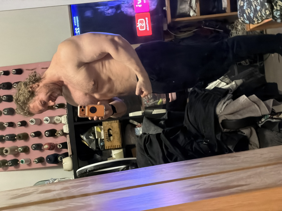 The image displays a muscular, light-skinned male individual taking a mirror selfie within an indoor residence in Saint Petersburg, Russia. The male, shirtless and wearing dark athletic pants, stands at a slight angle, smiling and presenting a partial flex of his left bicep. He holds an orange-cased smartphone with a circular black pop-socket in his right hand, aimed towards the mirror. His hair is light brown and wavy.
The background elements include:
- A light-colored wall adorned with numerous small, varied decorative objects, resembling miniature hats or thimbles, arranged in multiple horizontal rows.
- To the left of the subject, a black cube shelving unit holds a black plastic bag, a wooden crate with the Cyrillic inscription "НАРОД" (NAROD), and other indistinct items.
- To the right, a large flat-screen television is visible, displaying a dark screen with red and white graphic elements, including a stylized "TV" logo.
- The far left edge of the image shows a partial reflection of an orange curtain or drape with a vertical strip of purple light.
- The lower right section of the image shows a wooden surface, possibly a nightstand, cluttered with various objects, and a patterned textile, likely a bedspread.
- The floor beneath the subject is a light-toned hard surface.