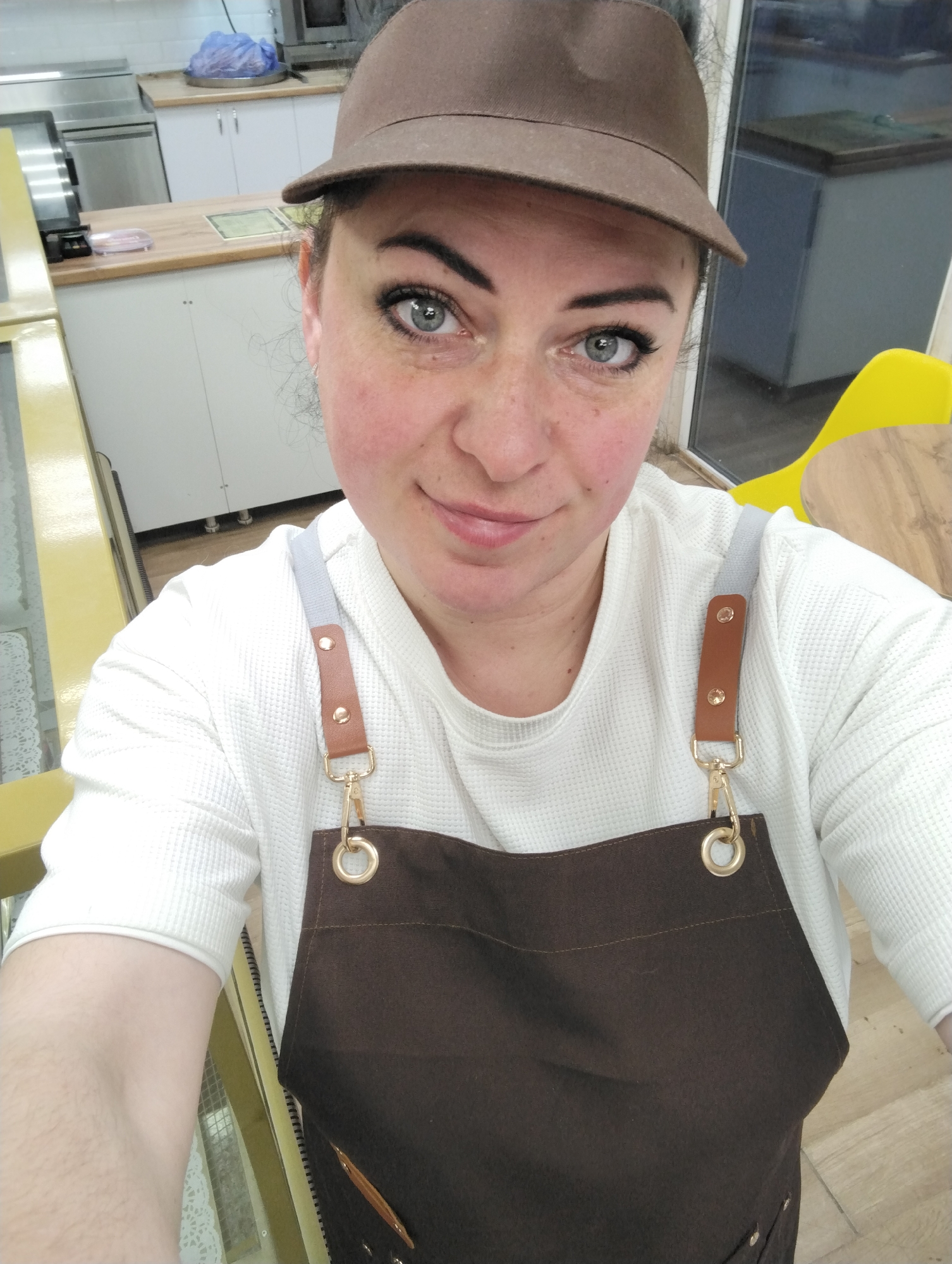A woman is taking a selfie indoors. She is wearing a brown cap and a brown apron over a white long-sleeved shirt. The apron has gray straps with brown leather and gold clasps. Behind her are kitchen cabinets, a microwave, and a yellow chair. Based on the available information, there is no verifiable evidence to confirm the location as Votkinsk, Russia.