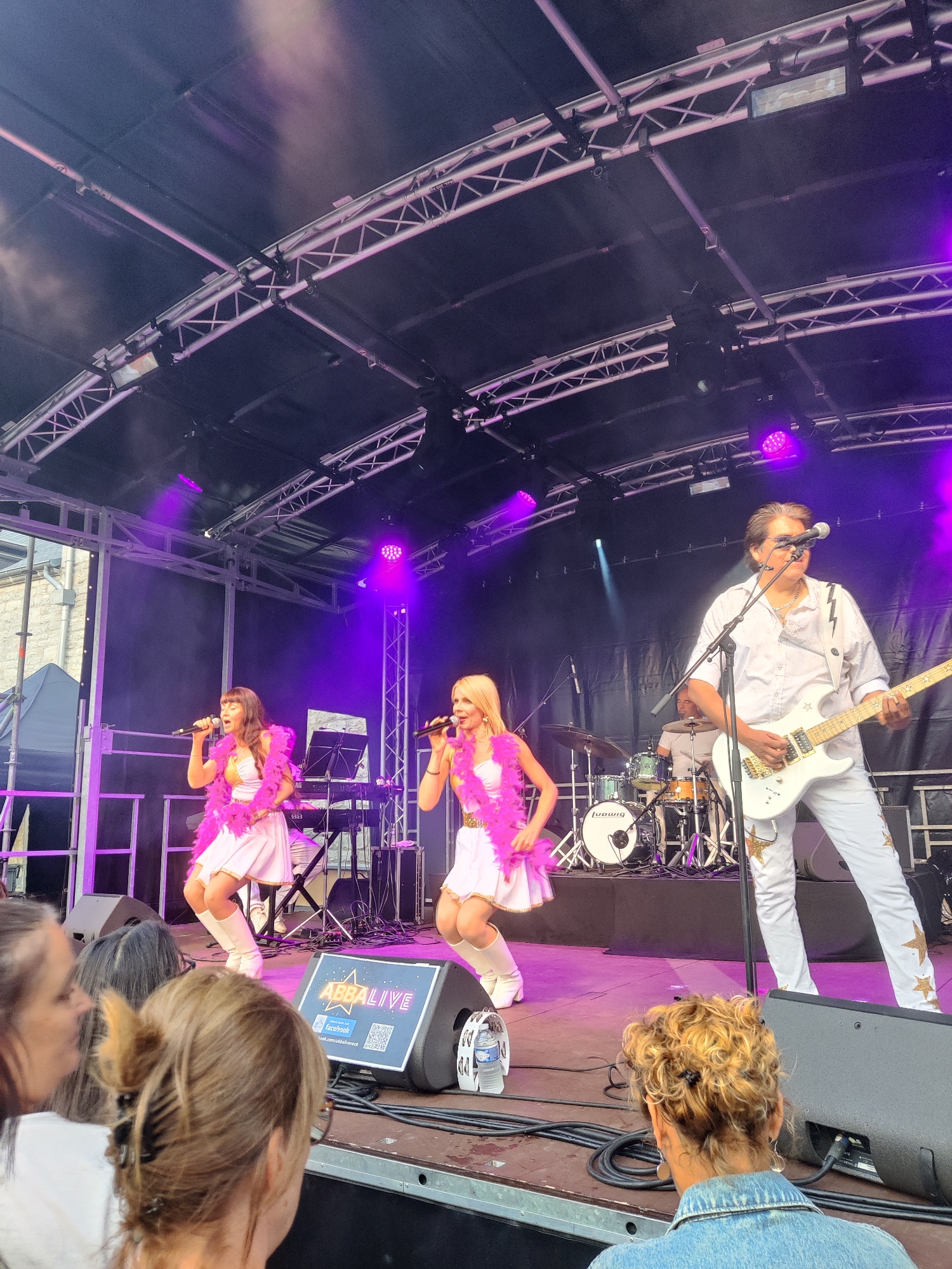 The image depicts an outdoor concert stage in Marche-en-Famenne, Belgium, where a band is performing. On stage, three people are visible: two women in matching white sequined dresses and pink feather boas, singing into microphones. The woman on the left has brown hair, and the woman on the right has blonde hair. A man in a white shirt and white pants adorned with gold stars is playing a white electric guitar. Behind the singers, a drum set with the brand name "Ludwig" is visible, and a keyboard is also present. The stage is set up with a dark canopy overhead, supported by metal trusses. Purple stage lighting illuminates the performers and creates an atmospheric effect with haze or smoke. In the foreground, the backs of several audience members are visible, looking towards the stage. A monitor speaker with the text "ABBAlive" and a QR code is placed at the edge of the stage. The backdrop suggests an urban or town setting with a stone building visible to the left of the stage. The overall scene captures a live musical performance under ambient lighting conditions.