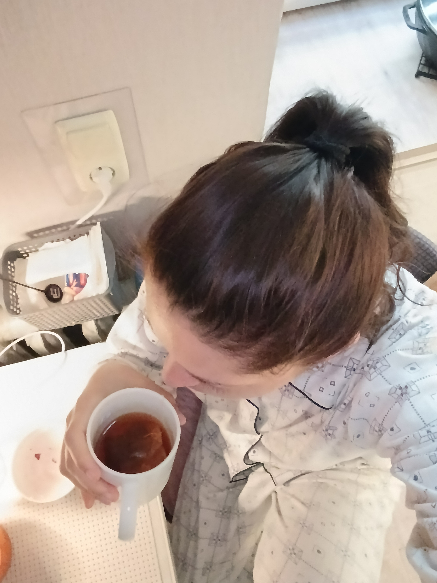 A person, with dark hair pulled into a ponytail, is holding a white mug of tea. The person is wearing pajamas with a geometric pattern. In the background, there is a white electrical outlet with a cord plugged into it, and a grey mesh basket containing white objects. The scene appears to be indoors, likely in a home. The image does not provide any information that would allow for specific location identification within Saratov, Russia.