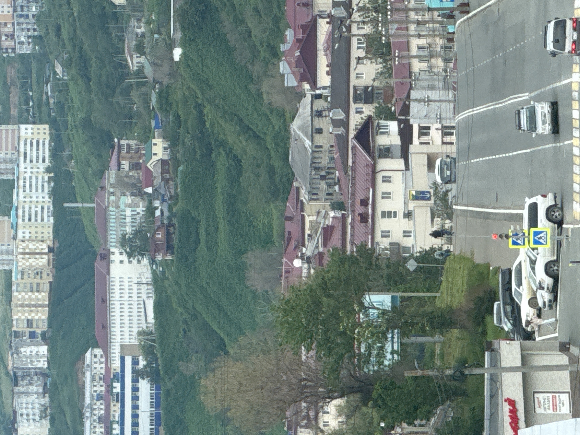 The image shows a view of the city of Kholmsk, Russia, with buildings and a street. The buildings are multi-story structures, some with flat roofs and others with pitched, red roofs. One large, light-colored building with many windows is prominent in the mid-ground. A green, hilly landscape with trees surrounds the urban area. A two-lane paved road runs through the foreground, with a pedestrian crossing. Several vehicles are present on the road, including a white SUV, a white sedan, a darker SUV, and two white vans. A person is walking across the pedestrian crossing. Traffic lights and road signs are visible. The sky is overcast.