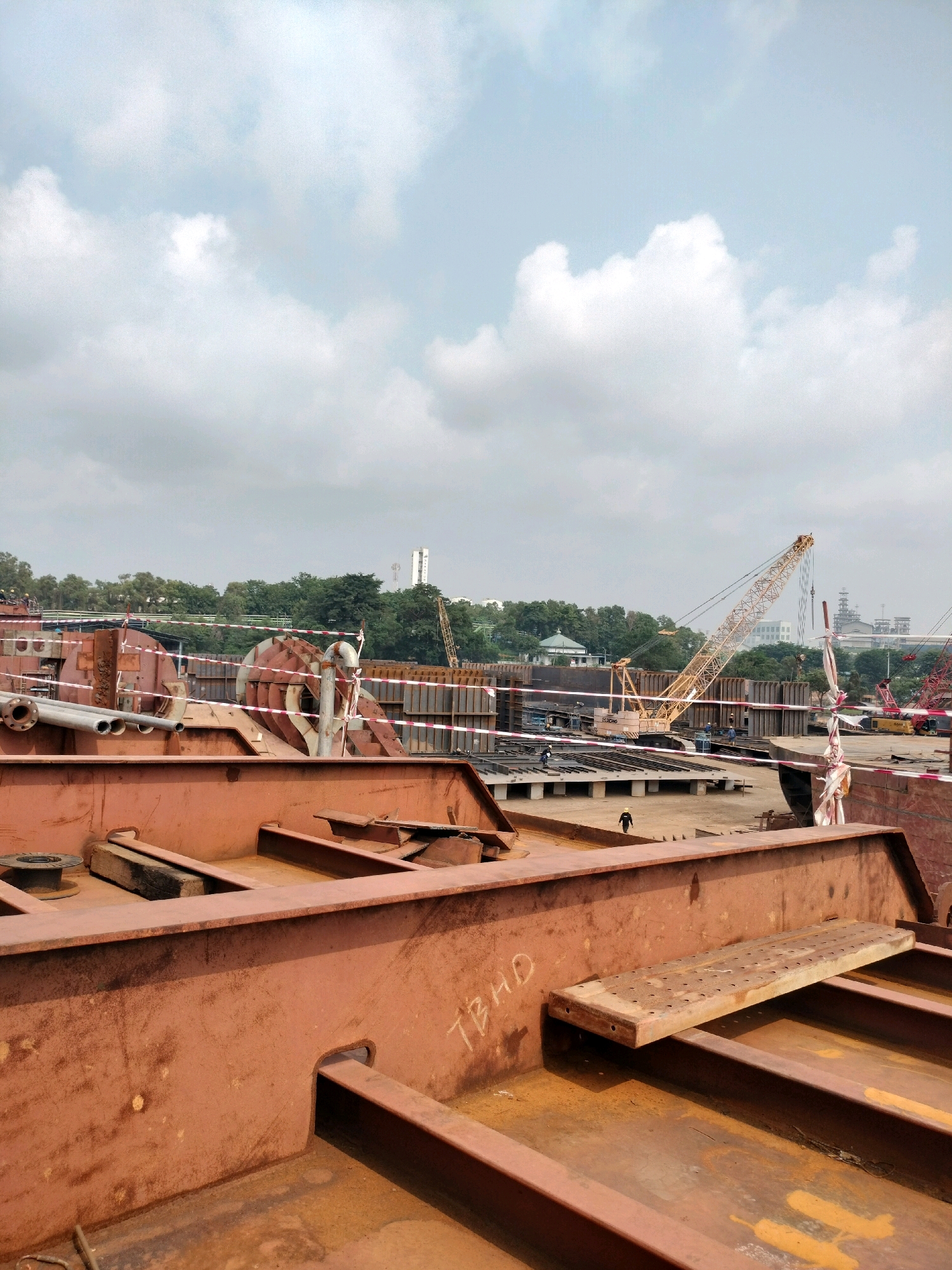 This image depicts a shipyard under construction in Batam, Indonesia. The foreground is dominated by large, rusty metal structures, likely components of a ship or vessel. In the mid-ground, a yellow crane is visible, along with other construction equipment and workers. Several unfinished sections of what appear to be ships are laid out, surrounded by construction materials and barriers. The background features a line of trees, buildings, and more industrial structures, suggesting the location is within an industrial area of Batam. The sky is overcast with clouds.