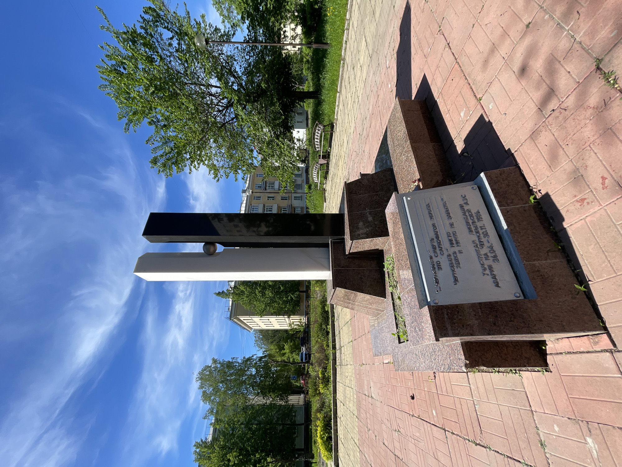 This image depicts a monument dedicated to the liquidators of the Chernobyl accident, situated within an urban park setting under a clear blue sky with cirrus clouds, likely in the city of Zheleznogorsk, Russia.
The central subject is a monument composed of two tall, rectangular, polished pillars of unequal height and width, one white/light-colored and one black/dark-colored, positioned side-by-side. A polished metallic sphere is embedded between the two pillars at roughly two-thirds of their height from the base. The pillars rest on a multi-tiered base made of polished red-brown granite. Attached to the front of this base is a rectangular metal plaque with an inscription in Russian.
The inscription reads:
"ЧЕРНОБЫЛЬ - СМЕРТЬ
ЭТО САРКОФАГОМ УКРЫТА"
(CHERNOBYL - DEATH
IT IS COVERED BY A SARCOPHAGUS)
"ЧЕРНОБЫЛЬ - НИКТО
ЗДЕСЬ И НИЧТО НЕ ЗАБЫТО"
(CHERNOBYL - NO ONE
HERE AND NOTHING IS FORGOTTEN)
"УЧАСТНИКАМ ЛИКВИДАЦИИ
АВАРИИ НА ЧЕРНОБЫЛЬСКОЙ АЭС
26.04. - 30.11. 1986"
(TO THE PARTICIPANTS OF THE LIQUIDATION
OF THE CHERNOBYL NPP ACCIDENT
26.04. - 30.11. 1986)
The monument is placed on a paved area consisting of reddish-brown and yellowish-beige rectangular pavers laid in a pattern. Small weeds are visible growing between some of the pavers and in the gaps around the monument's base. Strong shadows are cast by the monument onto the paving, indicating a high sun position.
Beyond the paved area, a strip of unkempt green grass separates the walkway from further landscaping. Two empty park benches with decorative metal frames and wooden slats are visible in the midground to the right of the monument. Several deciduous trees with full green foliage are scattered throughout the scene, some bordering the paved area and others further in the background. A lamppost with a spherical light fixture is visible on the right side of the image, behind the monument.
In the background, multi-story residential buildings, likely apartment blocks, with light-colored facades (yellowish-beige and white) and multiple windows, are visible through and above the trees. Some smaller vehicles are partially visible parked between buildings in the distance. The sky above is a vibrant blue with sparse, wispy white clouds. No people are present in the image.