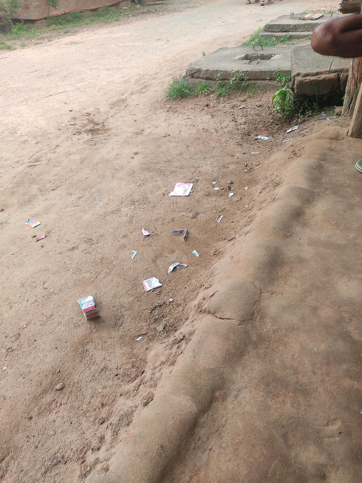 The image shows a close-up of a dusty, sandy area in MV 63, India. Scattered across the ground are several small, colorful papers, some appearing to be wrappers or candy packaging. A small stack of what looks like candy or small packets is visible in the lower left quadrant of the image. The ground itself is uneven, with some patches of dry earth and sand. In the background, parts of concrete structures and some greenery can be seen, suggesting an outdoor, possibly semi-urban or rural environment. The lighting suggests it is daytime.