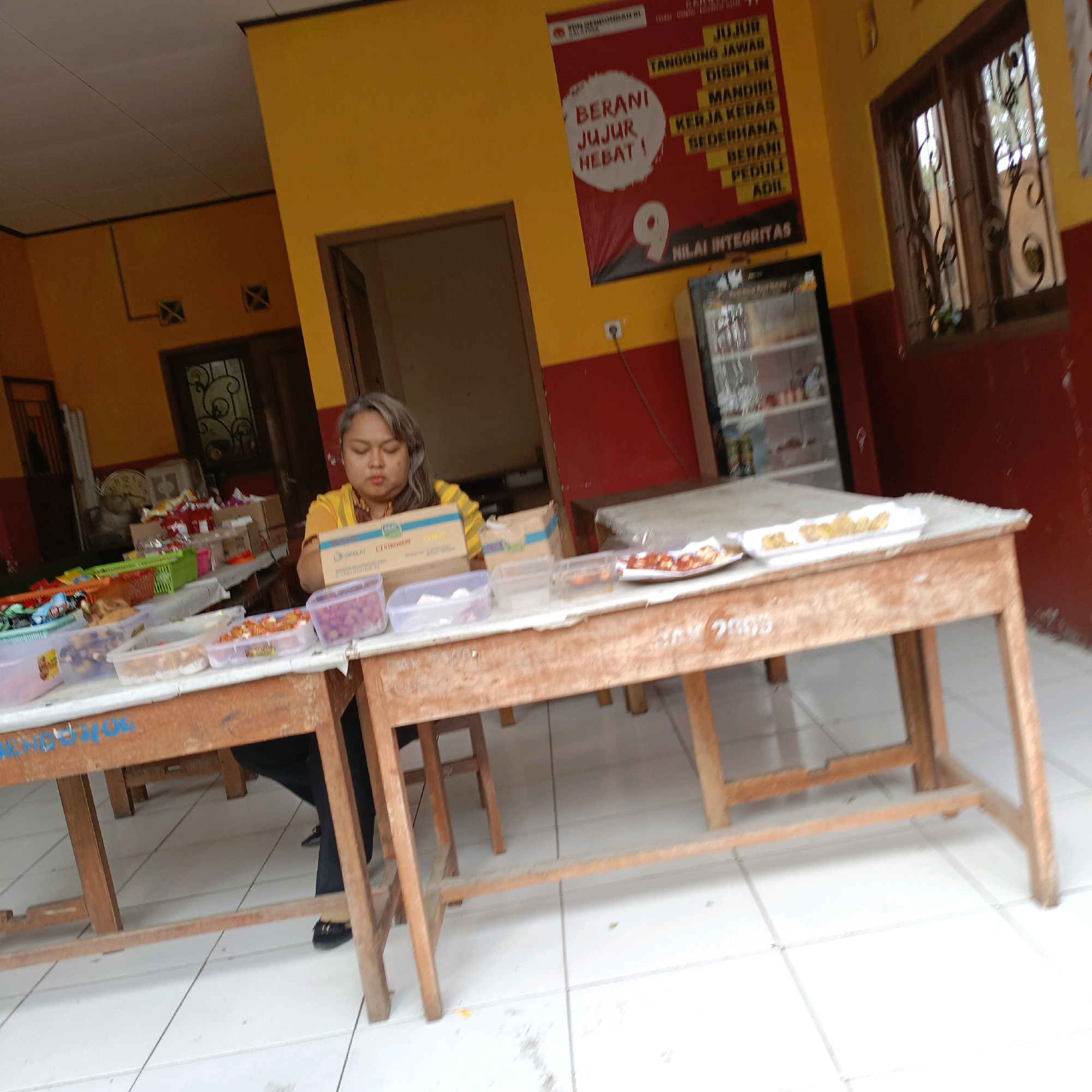 The image depicts an indoor scene, likely a cafeteria or a food stall, with a woman of Asian ethnicity, who appears to be in her late 30s or early 40s, sitting at a table. She has shoulder-length graying hair and is wearing a yellow top. In front of her, on the table, are several plastic containers filled with food, as well as a cardboard box.
The room's walls are painted in bright yellow and red. A large red poster with white and yellow text is visible on the left wall. To the right, a window with ornate metal bars and a glass pane is present, and further to the right, a refrigerator unit. Two wooden tables with checkered tablecloths are set up. The floor is tiled with white squares. The overall impression is of a functional, perhaps community-oriented, eating establishment in Salatiga, Indonesia.