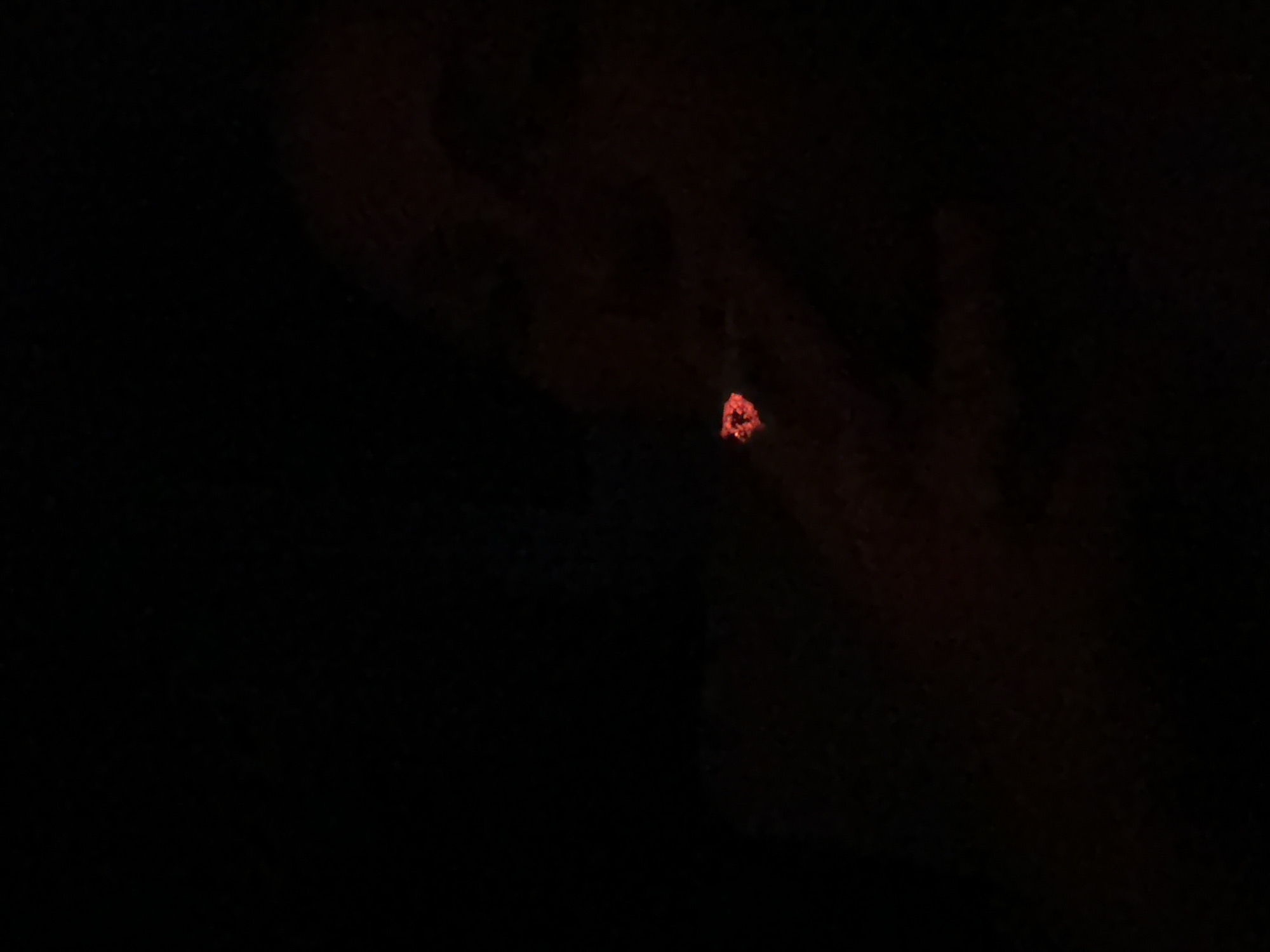 In a dimly lit environment, a close-up shot captures a glowing red ember at the tip of what appears to be a cigarette, held between fingers. The background is dark, with some indistinct shapes suggesting a person's hand and possibly clothing. The scene is devoid of any clear environmental or urban context, making it impossible to pinpoint a specific location within Kampot, Cambodia. The focus is solely on the small, brightly lit ember, which stands out starkly against the darkness. There are no other discernible subjects or objects in the frame.