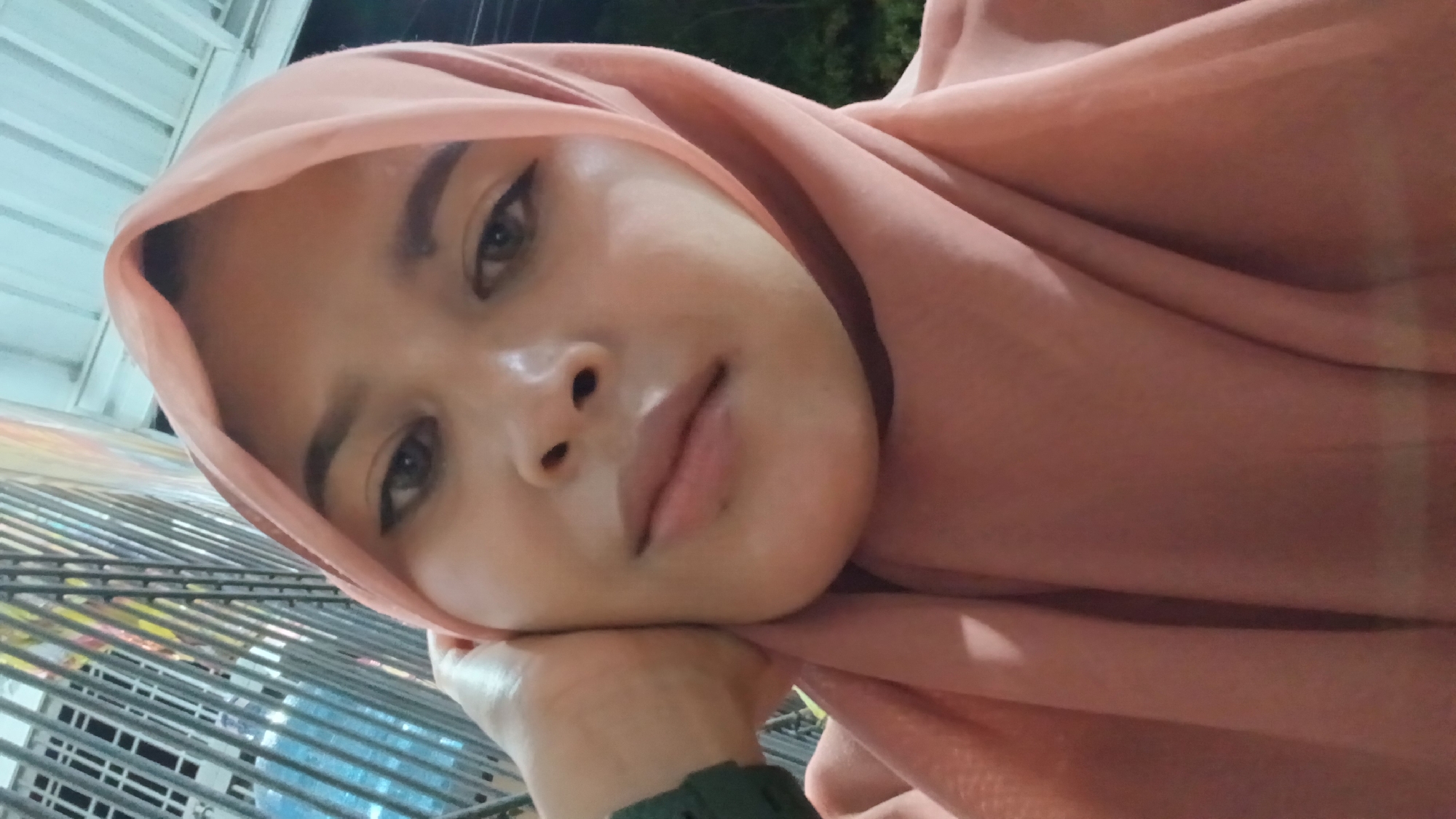 A close-up shot depicts a person wearing a peach-colored hijab, resting their chin on their hand. The person's face is angled towards the camera, revealing features such as dark eyebrows, winged eyeliner, and a slightly parted mouth. The background includes what appears to be a metal fence or structure, and glimpses of outdoor elements, possibly trees, are visible. The location is potentially within the city of Lhokseumawe, Indonesia.