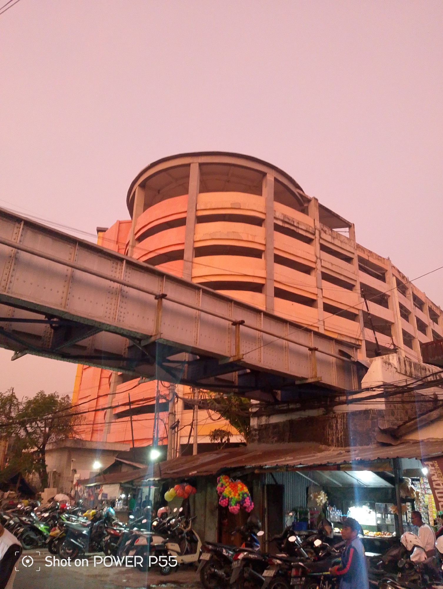 A multi-level parking structure with a rounded facade, painted orange and concrete, dominates the upper portion of the image. A metal skybridge, supported by visible beams and rivets, extends horizontally across the foreground, obscuring parts of the parking structure. Below the skybridge, a street scene unfolds. A dense arrangement of motorcycles is parked on the side of the road, with some appearing to be actively in use or waiting. A row of small open-air market stalls or shops lines the street, some with tin roofs, displaying various goods, including what appear to be balloons and bottled drinks. Several people are visible, including a person in the foreground wearing a blue and red jacket, and another person in a white shirt and head covering in one of the stalls. The sky has a pinkish, hazy hue, suggesting either dawn, dusk, or possibly pollution. The overall scene depicts a busy urban environment, likely a marketplace or commercial area, with a distinctive architectural element of the elevated parking garage and skybridge. Based on the provided information, this scene is located in the city of Surabaya, Indonesia.