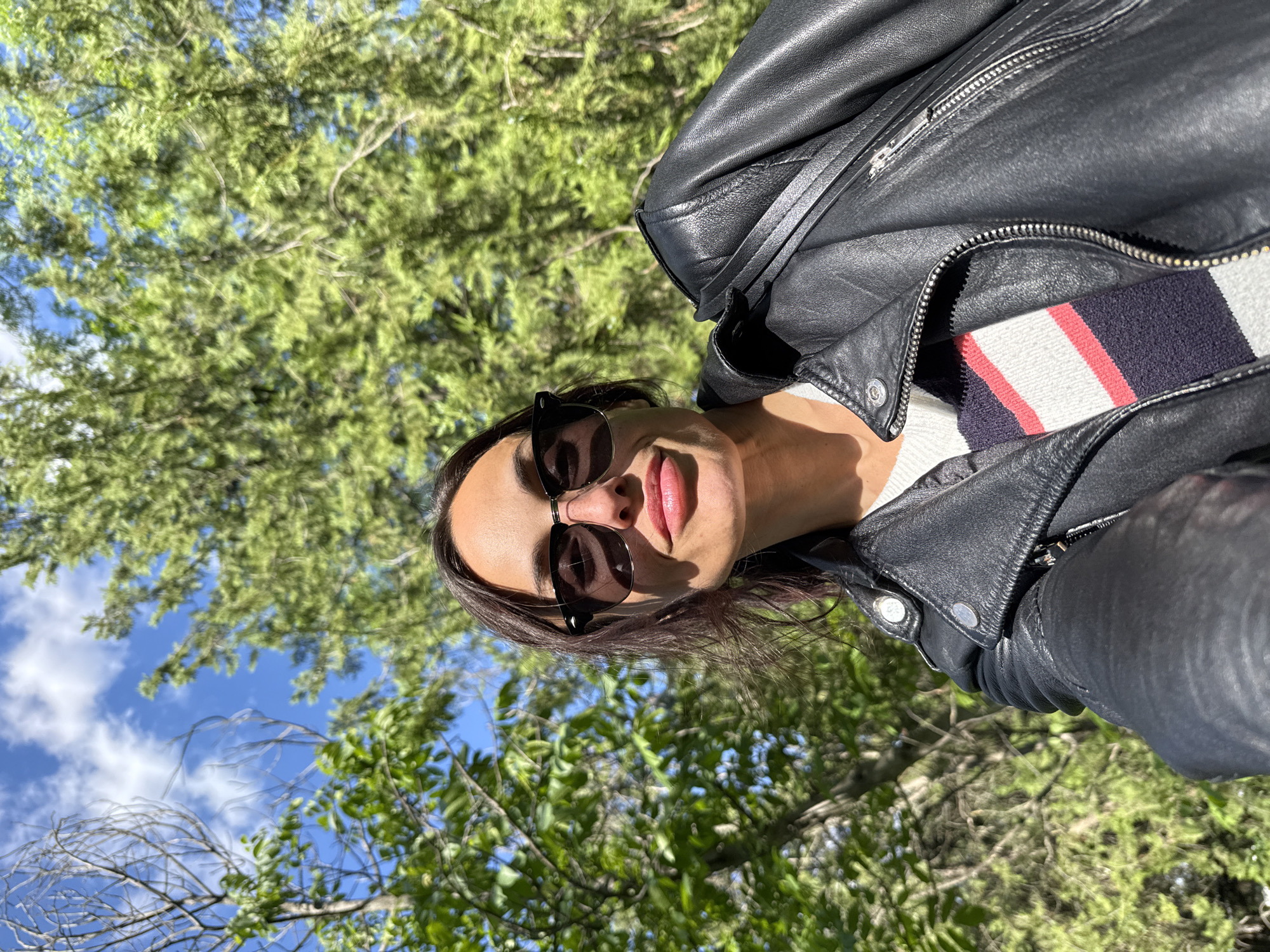 A medium, outdoor shot portrays a woman with sunglasses and dark hair, wearing a black leather jacket over a horizontally striped sweater. She is positioned in front of trees, with some branches and foliage partially obscuring the view. The background also shows glimpses of a blue sky. The photograph appears to have been taken from a low angle, looking upwards at the subject and the surrounding trees. The image does not offer enough context to confirm its location within the city of Pleven, Bulgaria.
