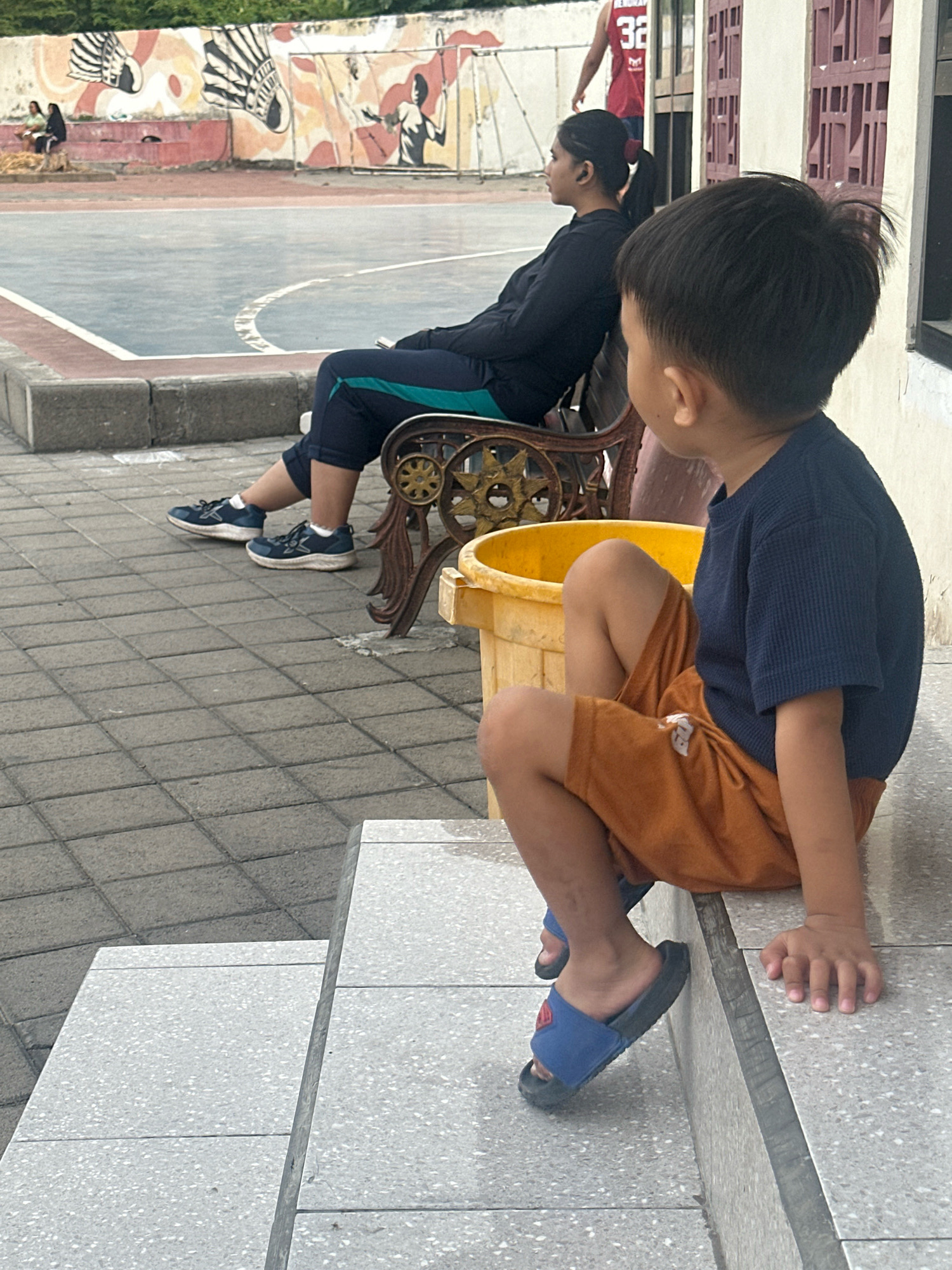 The image shows a young boy in the foreground, wearing a navy blue t-shirt and orange shorts, sitting on concrete steps. He is wearing blue slide sandals and his right hand is resting on the step beside him. In the background, a woman is sitting on a decorative metal bench, wearing a black long-sleeved shirt and dark pants with teal stripes. She is looking to her left. Behind her, a person in a red basketball jersey with the number 32 is visible. The setting appears to be an outdoor public space, possibly a park or sports court, with a painted mural on a wall in the background depicting badminton players and abstract designs. The ground is paved with grey interlocking tiles. This scene could be located in Mojokerto, Indonesia, given the context provided.