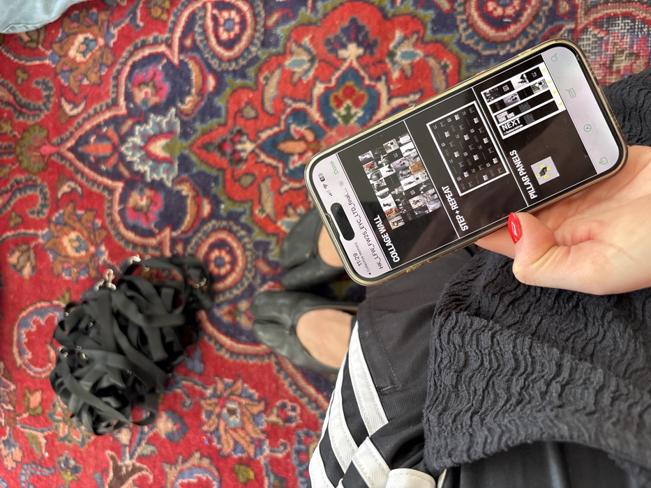 The scene is observed to be taking place in London, United Kingdom.
An overhead perspective captures the lower half of a person seated on a predominantly red patterned rug, which features intricate blue, green, and orange floral and paisley motifs. The person's bare feet are clad in black leather tabi-style ballet flats. Their left leg is partially visible, covered by a black garment with a broad white horizontal stripe, while their right leg and lap are draped in a black, crinkled, textured fabric. The person's right hand, with fingernails painted red, holds a gold-framed smartphone. The phone screen displays a digital document or presentation with the title "HK_LFW_FW25_EYC_LTD_final...", indicating content related to fashion week planning. Sections titled "COLLAGE WALL," "STEP + REPEAT," and "PILLAR PANELS" are visible, populated with various small fashion-related images and diagrams. The time "11:29" is shown in the upper left of the phone screen. To the upper left of the frame, a loose bundle of black fabric straps or ribbons, some with small metallic silver findings, rests on the rug.