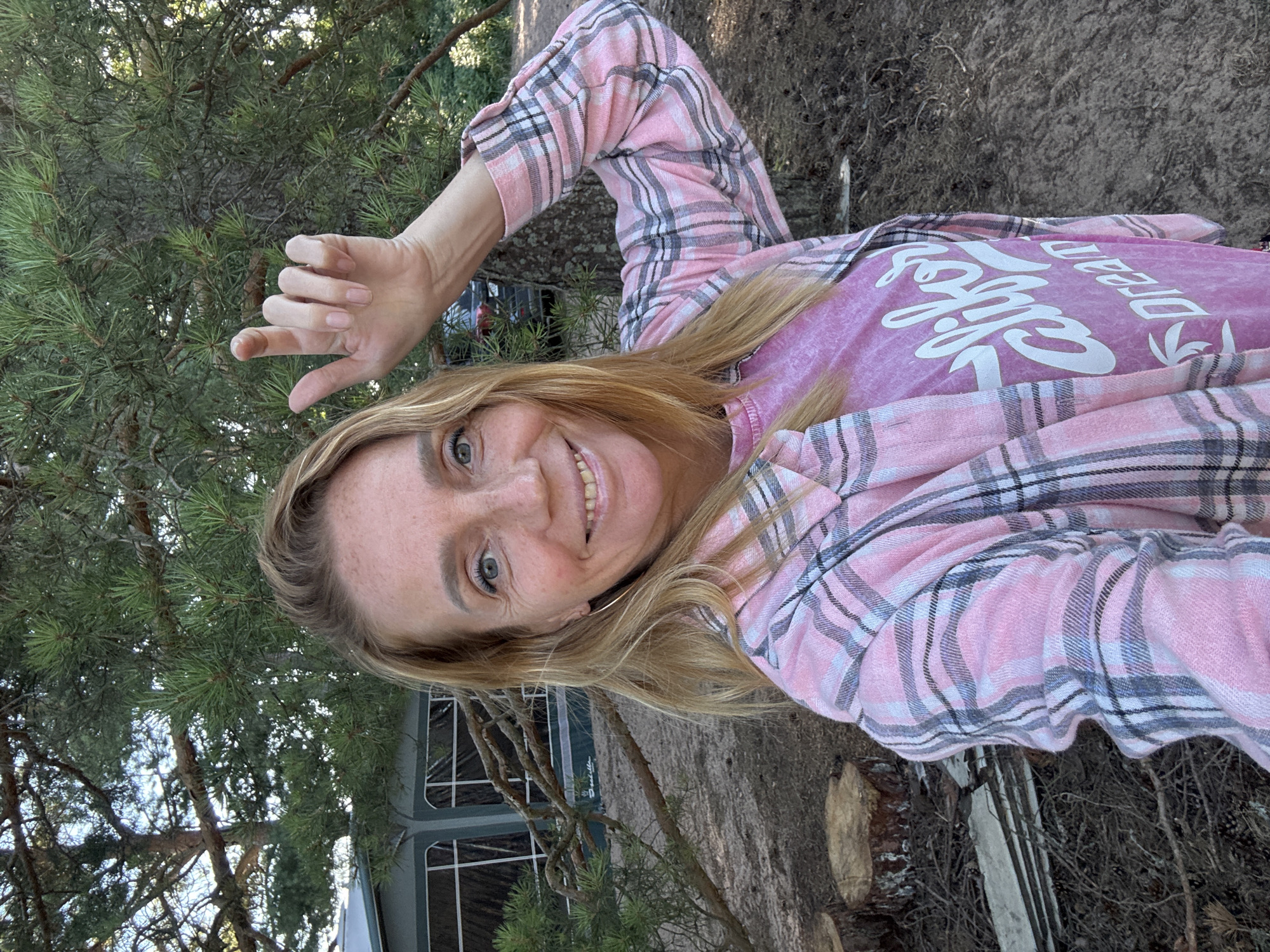 A middle-aged Caucasian woman with blonde hair and freckles is taking a selfie outdoors. She is wearing a pink and grey plaid shirt over a pink t-shirt with white lettering that reads "California Dream". Her right hand is raised, with her index finger pointing upwards. The background shows trees, a large caravan or mobile home with windows, and several tree stumps on the ground. The lighting suggests it is daytime, possibly late afternoon. The location appears to be a natural or semi-natural environment, possibly a campsite or a wooded area near housing. It is not possible to determine the specific location within Zelenaya Roshcha from the image alone.