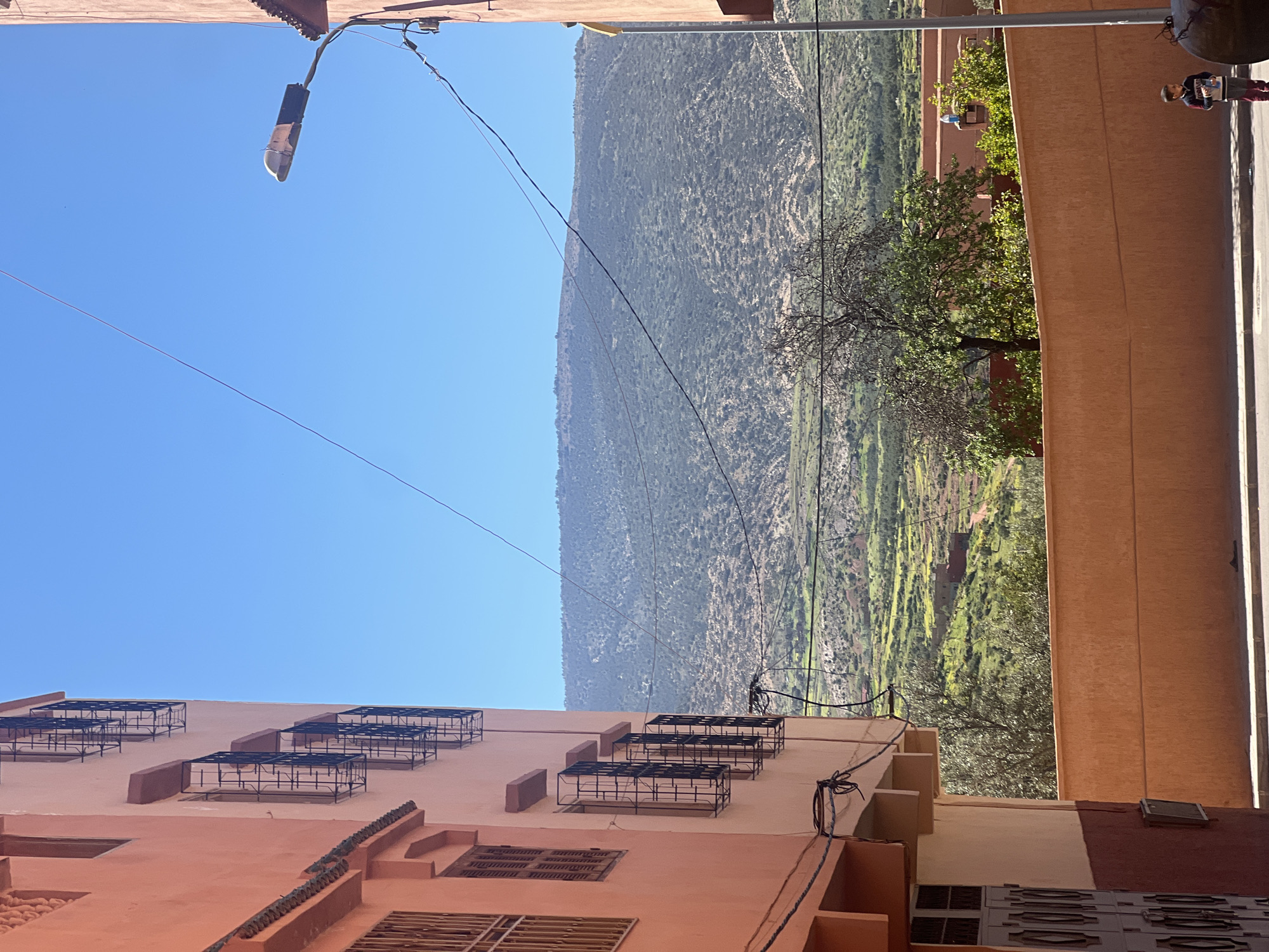 The image depicts a street view in Ouaouizert, Morocco, featuring buildings, mountains, and utility poles. The architecture consists of multi-story structures with peach-colored facades and dark window frames. The mountain range is visible in the background, with vegetation covering its slopes. A street light attached to a pole with cables is visible against a clear blue sky. A person is visible on a street sidewalk in the top right corner of the image.