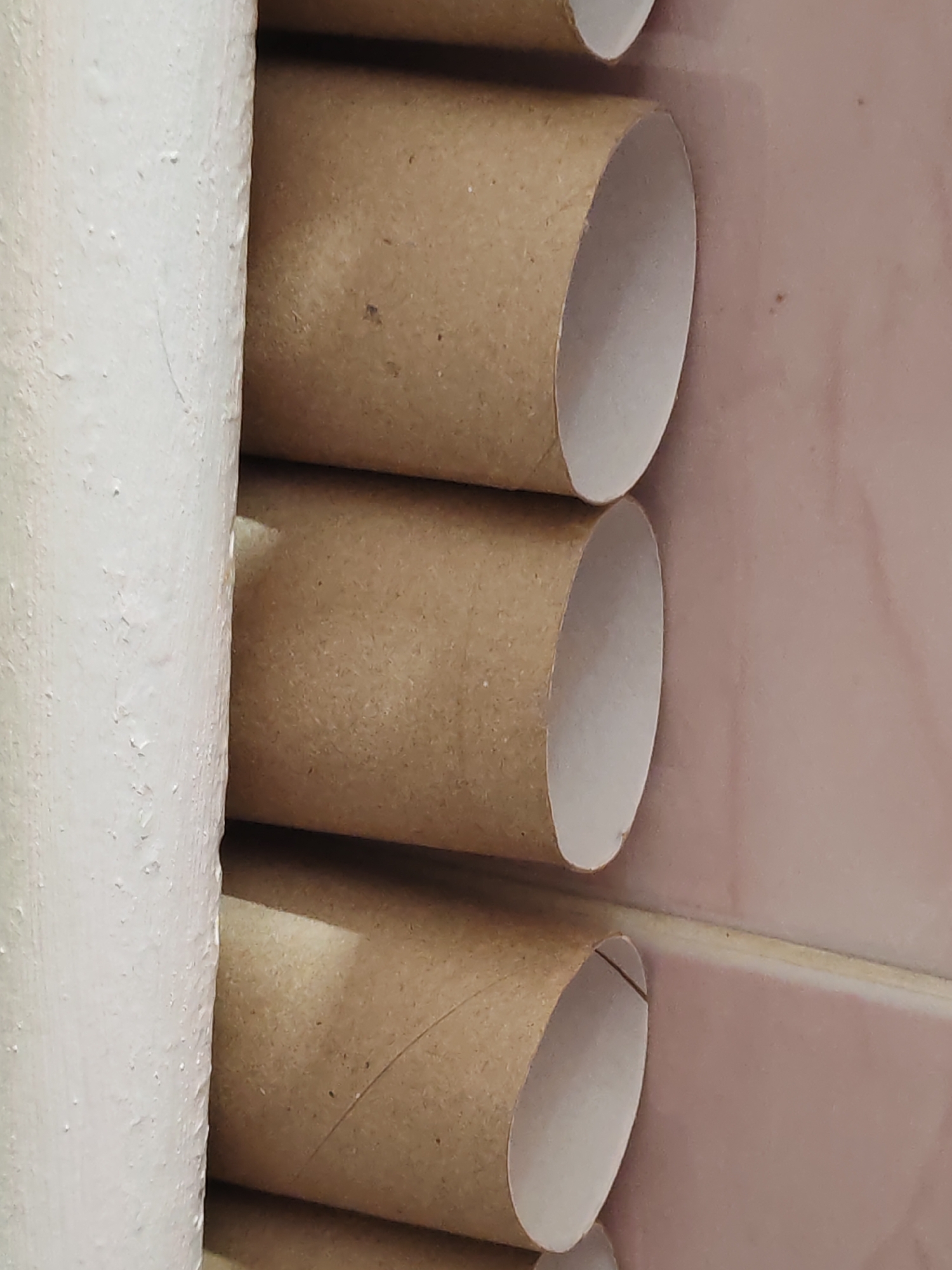 The image shows a close-up of stacked cardboard toilet paper rolls next to a white painted cylindrical object. The background reveals a pale pink tiled surface. No information is provided about the location in a city.