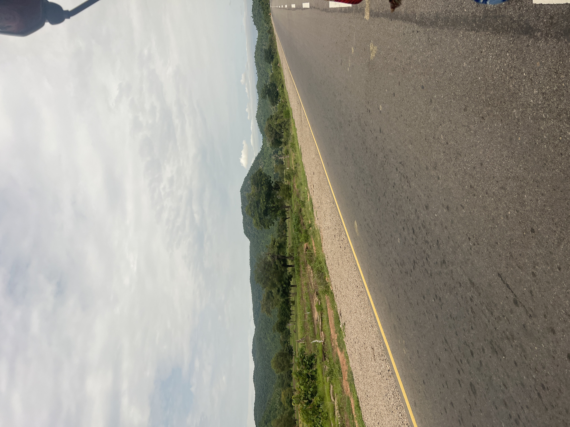 The image depicts a road in Minda, Nigeria, with a view of lush green hills and a cloudy sky. The road is asphalt with a yellow line marking the edge. The scene is likely captured during the day, given the bright sky. There are trees and vegetation along the side of the road, suggesting a rural or semi-rural setting. The perspective indicates the photo might have been taken from a moving vehicle or by someone standing by the roadside. The cloudy sky suggests it might be a humid or overcast day. There are no visible people or specific objects that stand out, other than the road itself and the natural landscape. The overall mood is tranquil, focusing on the vastness of the landscape and the open road.