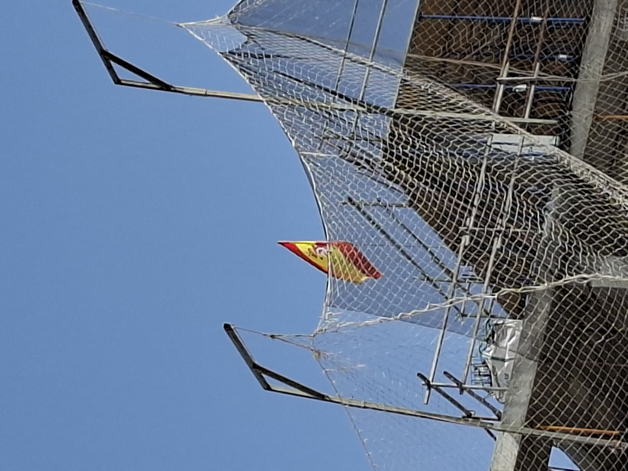 The image shows a Spanish flag flying from a construction site in Torrevieja, Spain. The flag is red and yellow with an emblem in the center, and it is attached to a metal pole extending from a scaffolding structure covered with netting. The background is a clear blue sky. The scene depicts ongoing construction with visible materials and equipment.