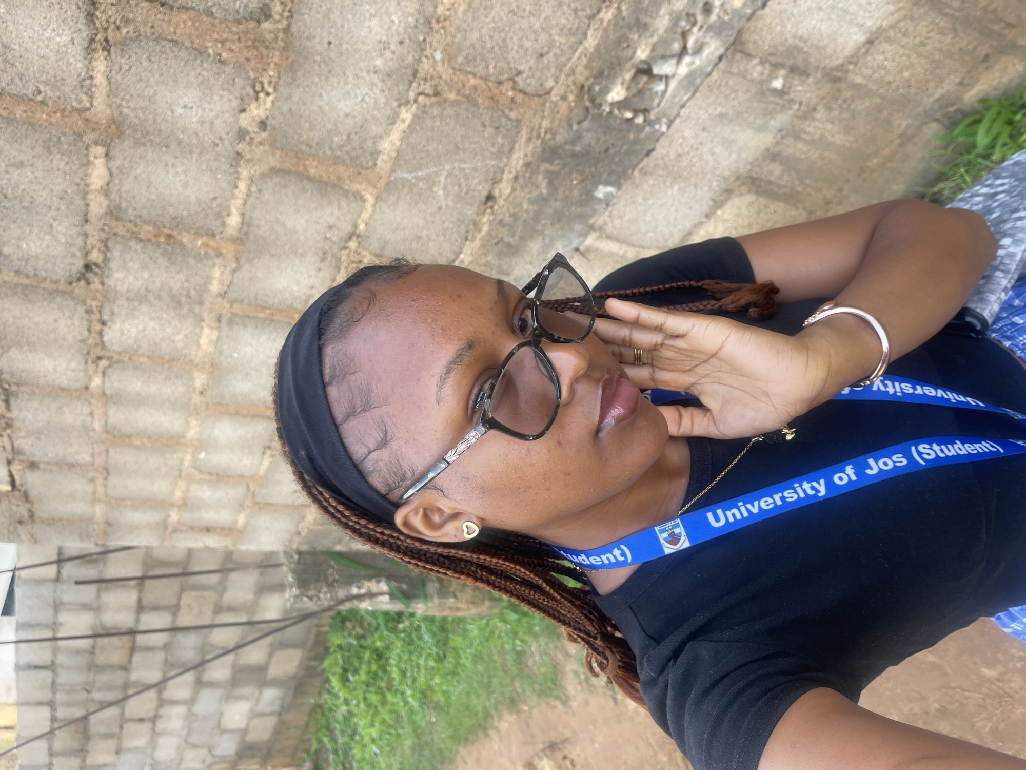 A medium shot captures a young woman with braided hair, wearing a black t-shirt, glasses, and a University of Jos (Student) lanyard. She is positioned in front of a rough-hewn brick wall, with some greenery and a dirt path visible to her left. The woman's right hand is touching her cheek. The scene is outdoors, likely in an urban or semi-urban setting, possibly within the vicinity of Rafin Par, Nigeria, suggested by the visible brick construction and vegetation.