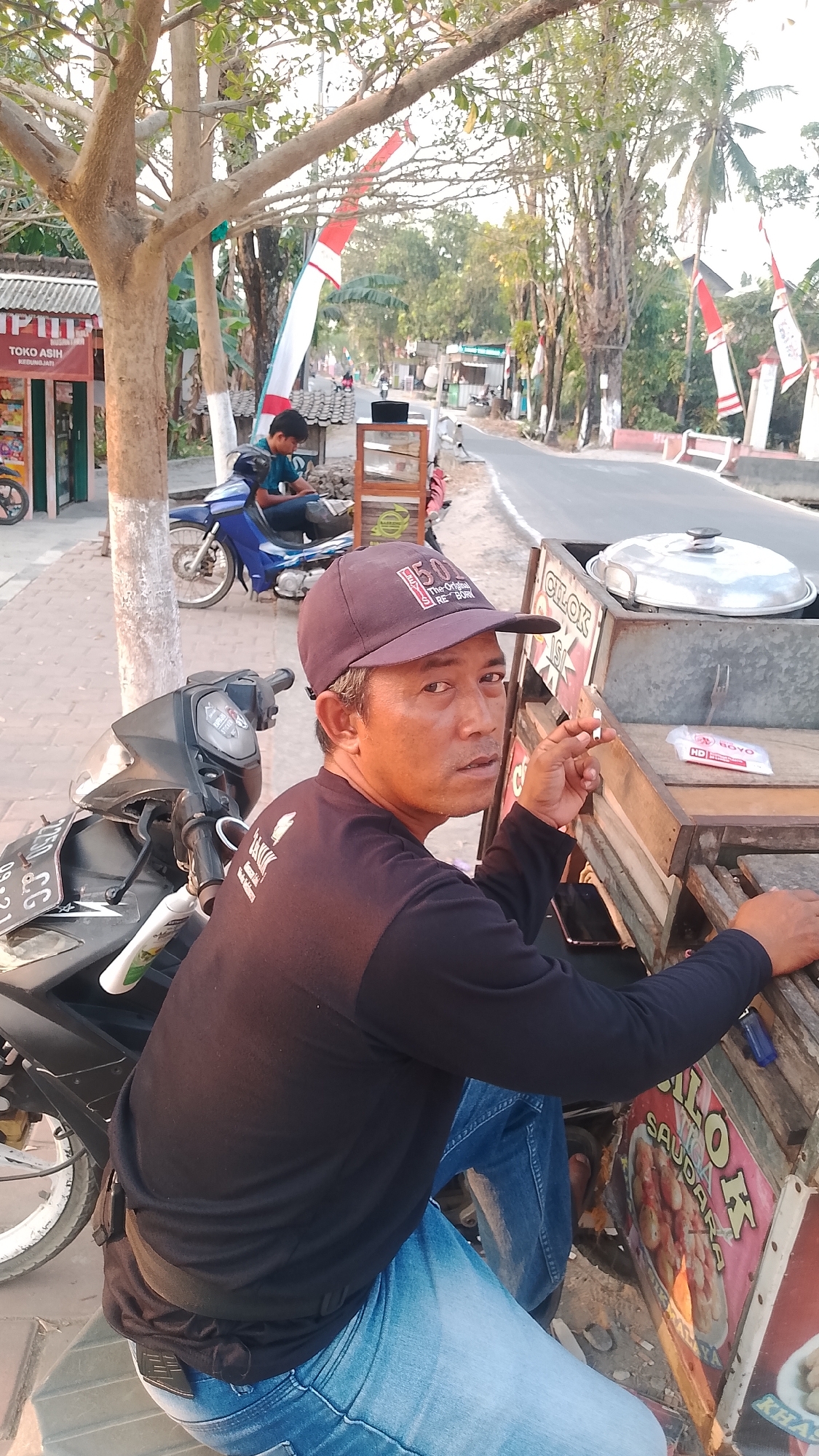 In Karanglangu, Indonesia, a man with a baseball cap and a black long-sleeved shirt sits on a motorcycle parked on a paved road. He is holding a cigarette and appears to be looking at the camera. Beside him is a wooden cart with various items, including a pot with a lid and a sign advertising "CILOK SAUDARA." In the background, a blue motorcycle is parked, and a person wearing a blue shirt sits on it. The scene is outdoors, with trees and Indonesian flags visible along the roadside. The lighting suggests it is daytime, possibly late afternoon due to the warm tones.