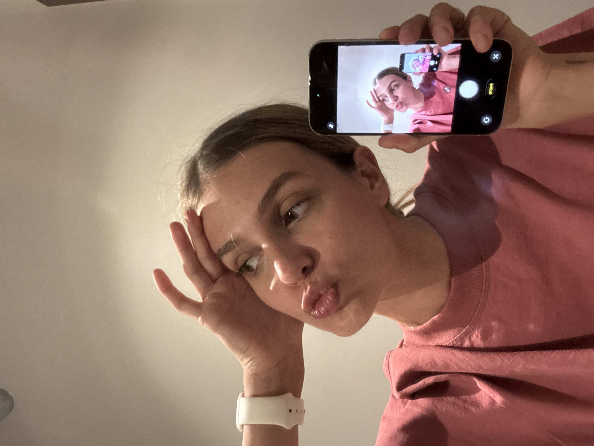 A woman is holding a smartphone in her right hand, taking a picture of herself in the phone's front-facing camera. The woman is wearing a salmon-colored shirt. Her left hand is near her forehead, and she is making a kissing face. She is wearing a white smartwatch. The background is a plain white or light-colored wall. The location is Unknown, Unknown.