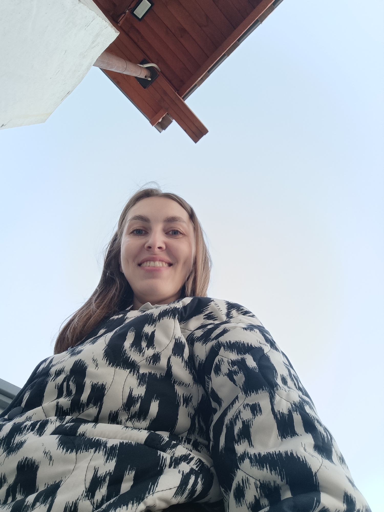 A low-angle, upward-facing selfie of a woman, possibly in her late 20s or early 30s, with long, light brown hair and a slightly tilted head. She is smiling and wearing a black and white patterned, quilted jacket. Above her is a bright, pale blue sky, with a portion of a building visible at the top of the frame. The building appears to have a white exterior wall and a brown wooden roof with a gutter and a light fixture. There is insufficient information to definitively confirm the location as Chaikovsky, Russia.