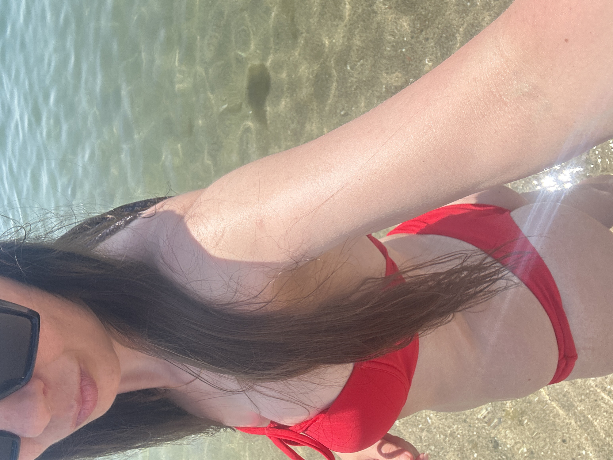 The image features a person wearing a red bikini, partially submerged in clear, shallow water. The water is transparent, revealing a sandy bottom with sunlight dappling through the surface. The person's skin, arm, and part of their face are visible. They are wearing dark sunglasses. Long, dark hair flows around their shoulders and across their body. The scene appears to be a beach or a shallow body of water, bathed in bright sunlight, suggesting a warm, sunny day. The overall impression is one of leisure and relaxation in an outdoor environment. The location context is Barcelona, Spain, but this is not visually apparent from the image itself. There is no visible text in the image.
