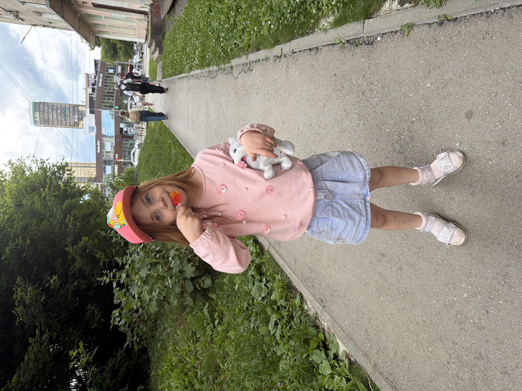 A young girl with long, reddish-brown hair and blue eyes, wearing a pink baseball cap with a cartoon character, a pink long-sleeved shirt adorned with fabric flowers and pearls, and a light blue denim skirt, stands on a paved sidewalk. She is holding a red lollipop to her mouth with her right hand and a small, white stuffed animal in her left arm. She is wearing white socks and light-colored sandals. The sidewalk is bordered by a strip of green grass and plants on the left and a concrete curb with more grass and small white flowers on the right. In the background, several people are walking on the sidewalk, and beyond them are multi-story buildings, including a tall, modern skyscraper. This scene is likely located in the city of Perm, Russia, given the architectural style of the buildings and the overall urban environment. The sky is overcast with clouds.