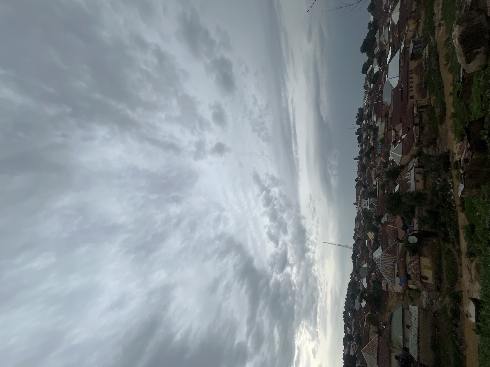 The image shows a dense residential area in the city of Jos, Nigeria, under a cloudy sky. The buildings are predominantly single-story structures with corrugated metal roofs, some of which appear aged and rusted. The houses are clustered together on a hillside, with narrow pathways or unpaved roads winding between them. Vegetation, including trees and grass, is visible in various parts of the scene, some appearing lush and green, while other areas show signs of dryness or disturbed earth, possibly due to recent rain.
Prominent in the background, a tall tower, likely a telecommunications mast, stands out against the overcast sky. There are no visible people or specific actions taking place. The overall impression is of a typical, densely populated urban settlement in a somewhat hilly terrain, under a weather condition that suggests impending or recent rainfall. The lighting is diffused due to the cloud cover, creating a muted color palette dominated by earthy tones of the buildings and the greens of the vegetation, contrasted by the grey of the sky.