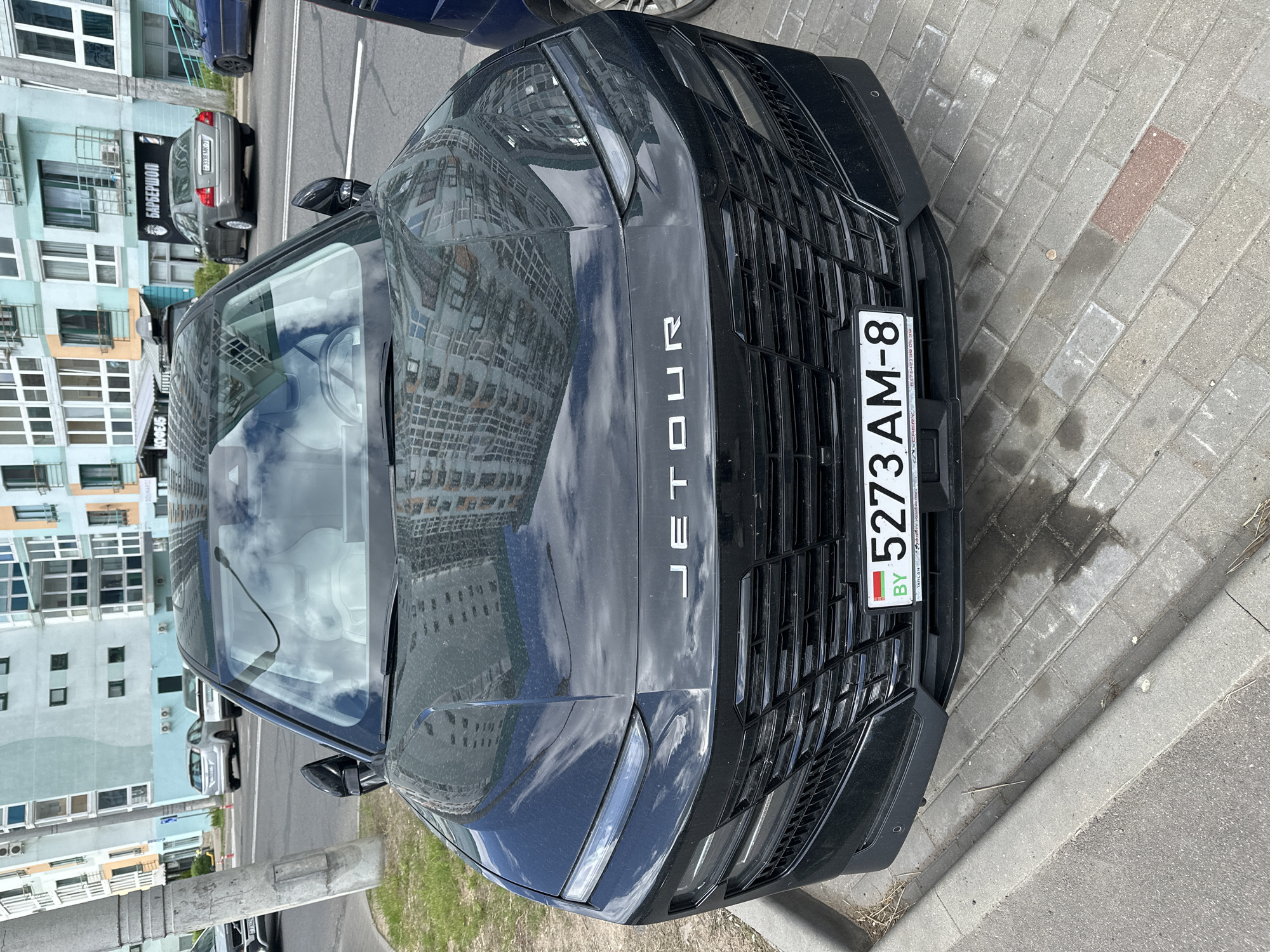 The image shows the front of a black Jetour vehicle parked on a brick sidewalk in Minsk, Belarus. The license plate number is 5273 AM-8. The reflection in the car hood reveals a nearby building. Across the street, other cars are parked near a building with a sign reading "БАРБЕРШОП". The sky is partially visible due to the reflection in the vehicle.