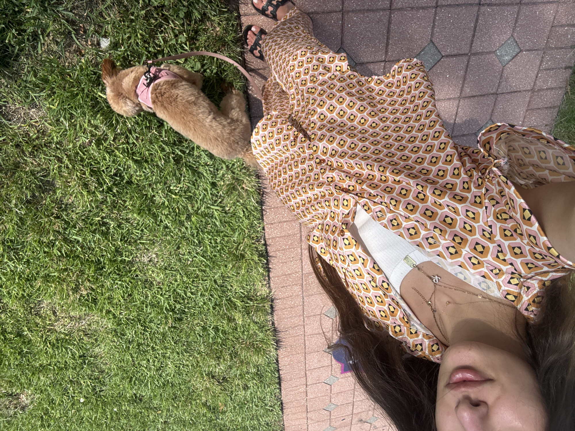 A woman with long brown hair is captured from the chest down, walking on a brick pathway next to a patch of green grass in Boca Raton, Florida, United States. She is wearing a pink, orange, and yellow geometric patterned pajama set, a white tank top, and black sandals. Around her neck, she has two delicate necklaces. She is holding a leash attached to a small, light brown dog wearing a pink harness. The dog is walking on the grass, sniffing the ground, while the woman walks on the adjacent brick path. The sun is casting shadows, indicating it is daytime.