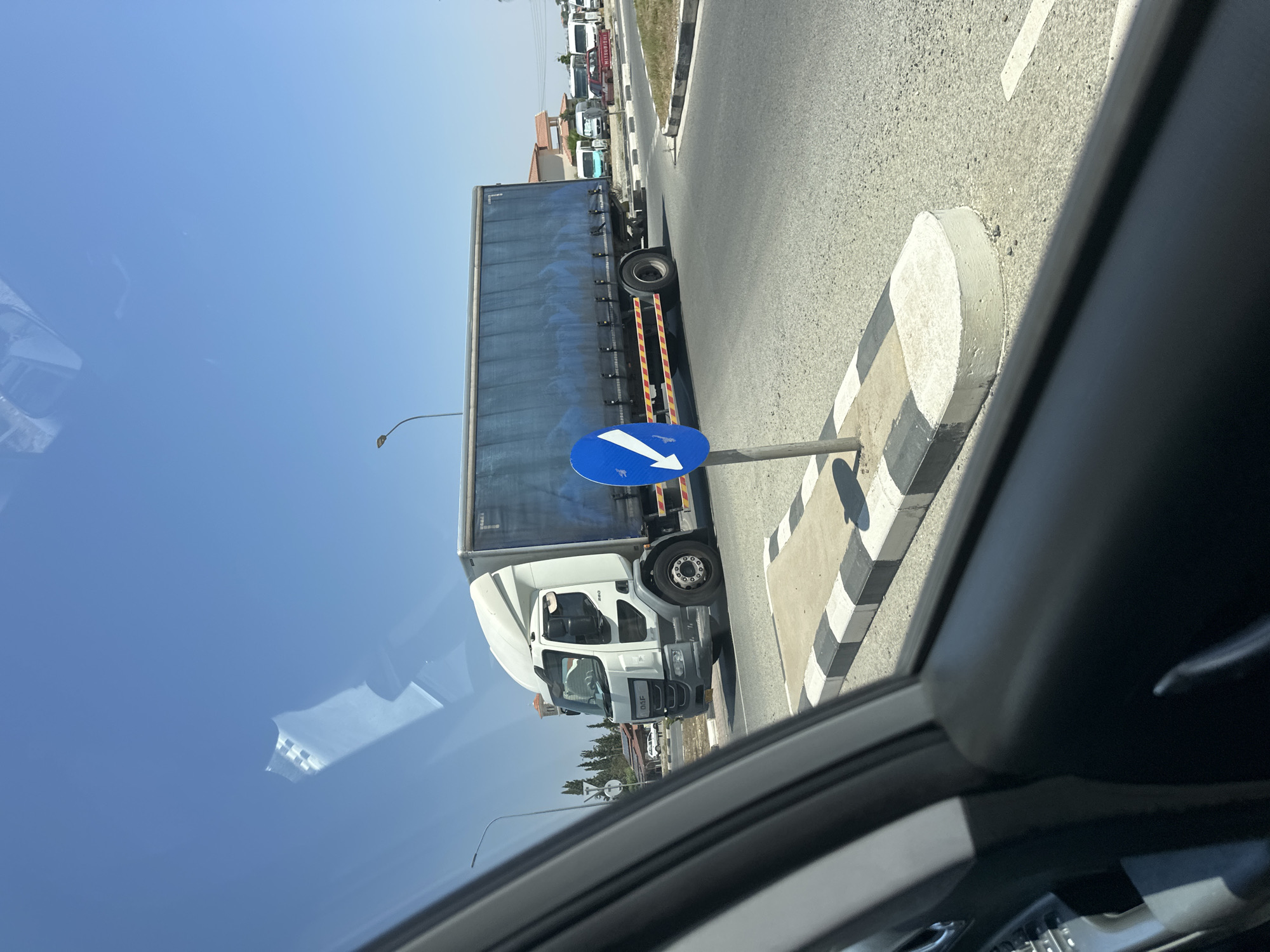 The image shows a white DAF truck with a blue tarp-covered trailer driving on a road under a clear blue sky. A blue circular traffic sign with a white arrow pointing down and to the left is visible in the foreground, next to a striped concrete median. The truck is in motion, and reflections are visible on the truck's windows and the surrounding glass surfaces. In the background, there are buildings, trees, and other vehicles, including vans and a red Mitsubishi truck. The scene appears to be a street intersection in a city.