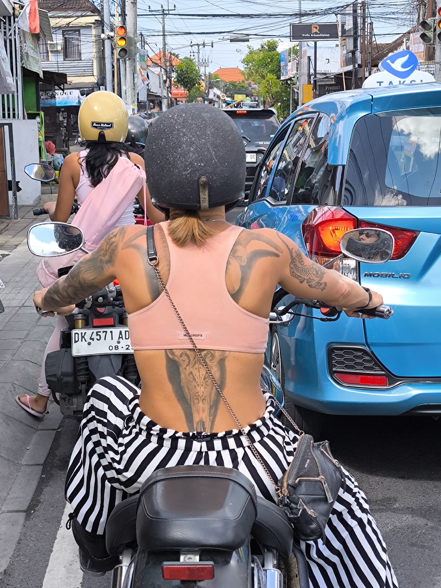 Captured from behind, two women on motorcycles are paused in traffic on a street in Canggu, Indonesia. The woman in the foreground wears a speckled black helmet, a peach-colored tank top, and black-and-white striped pants. Prominent tattoos are visible on her arms and back. A small black purse hangs on her side. Her companion wears a yellow helmet and a light pink outfit. A light-blue taxi is next to the woman in the foreground. A traffic light is visible. Buildings line the street. A road sign with a blue bird on it is visible above the taxi. The number plate on the woman's motorbike is DK 4571 AD, with the numbers 08.2 written below it.