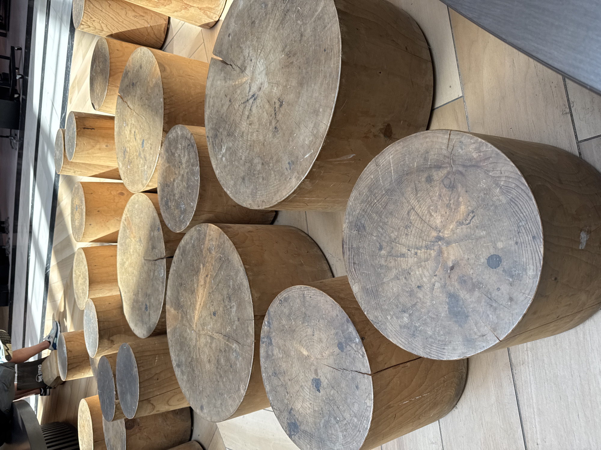 The image shows a vertical view of a wall constructed from large, horizontal wooden logs. The logs are of varying diameters and are stacked closely together, creating a textured, naturalistic surface. The grain of the wood is visible, with some logs showing prominent rings and a weathered appearance. The light source appears to be from the left, casting shadows and highlighting the texture of the wood.
In the lower left corner, a person's legs and feet are visible, wearing casual clothing including shorts and sneakers. The person is seated, with their legs extended towards the log wall. The background consists of a tiled floor and part of a dark structure, possibly furniture or a wall, to the left. The overall setting appears to be indoors, possibly in a public space or a modern building with natural material elements, given the clean tiling and the construction of the log wall. There are no overt indications of time of day or weather, but the lighting suggests it is daytime. The mood is relaxed and casual, due to the presence of the person and the natural, earthy tones of the wood. There is faint text visible on the person's shorts, which appears to read "THE STING". The location context of Leidschendam, Netherlands, implies this could be a public seating area or decorative feature in a shopping center, park, or other urban space.